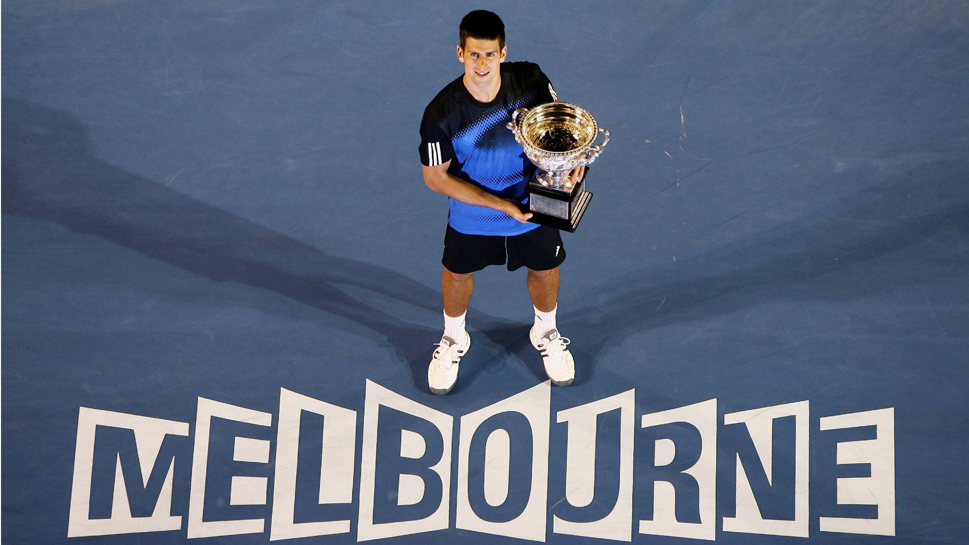 Australian Open 桌布 壁紙 — 網球冠軍諾瓦克·喬科維奇在藍色硬地球場上勝利地站立，手持閃亮的銀色獎盃，下方有大型白色字母寫著「MELBOURNE」。