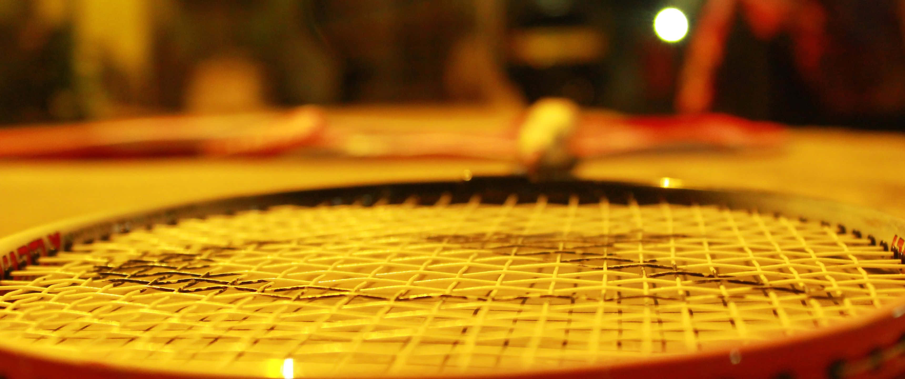 3440x1440p Badminton 桌布 壁紙 — 網球拍線床在金色室內燈光下溫暖發光的特寫，背景有一個模糊的球。