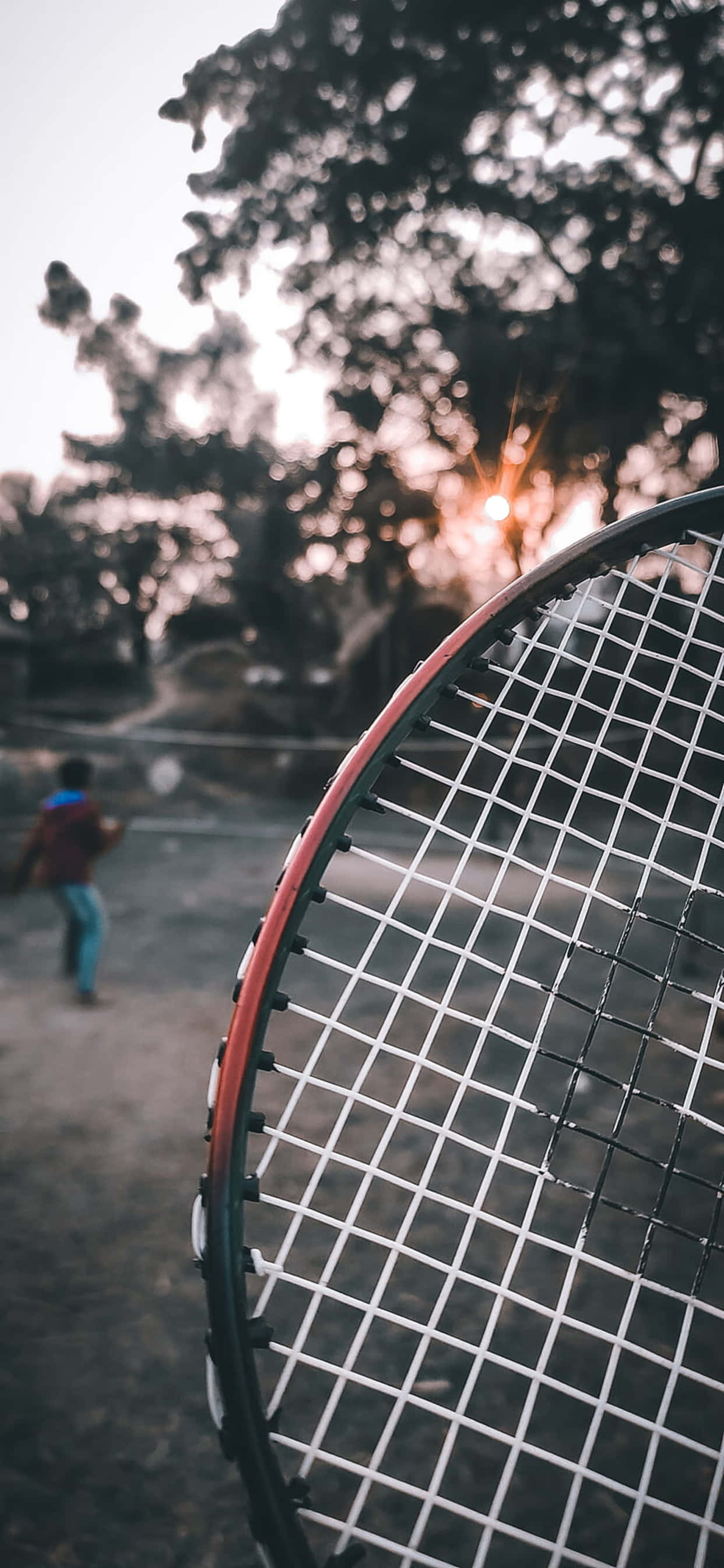 iPhone Xs Max Badminton 桌布 壁紙 — 網球拍頭的近距離特寫，背景有模糊的人物在金色日落時分打球。