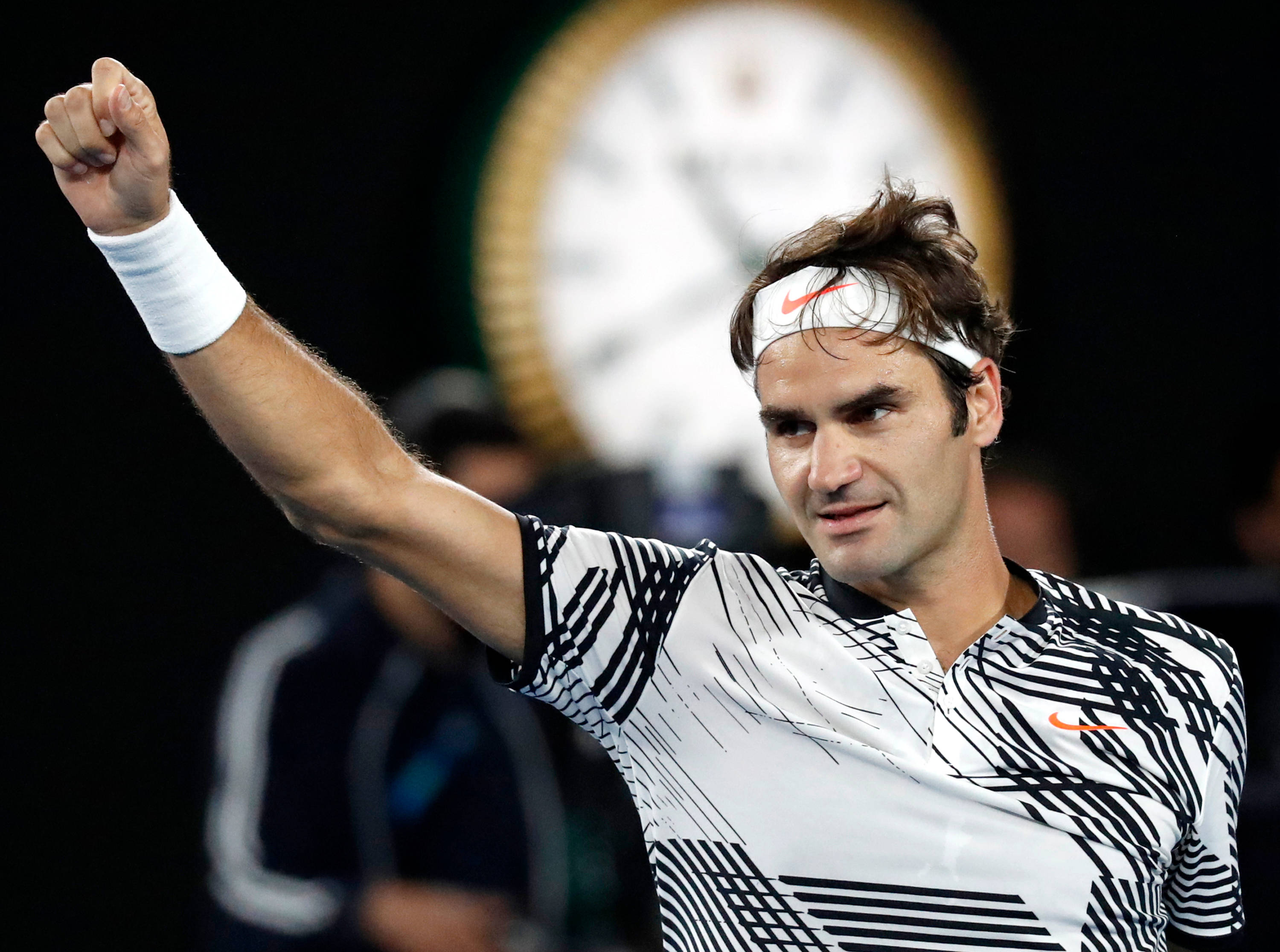 roger 桌布 壁紙 — 網球選手在明亮的體育場燈光下以勝利姿勢舉起緊握的拳頭，背景有模糊的時鐘。