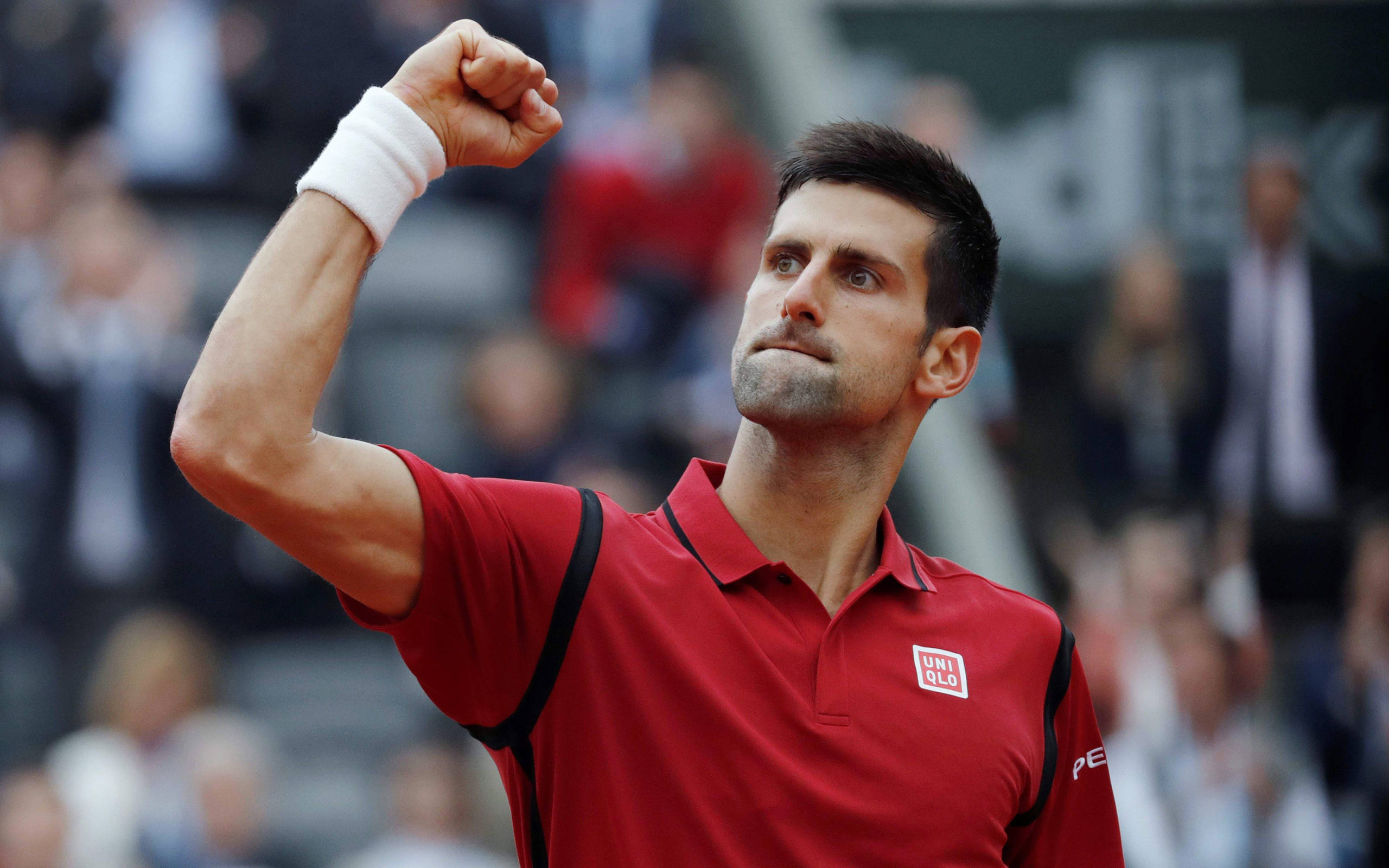 Novak 桌布 壁紙 — 網球選手諾瓦克·喬科維奇穿著紅色襯衫在紅土球場上握拳慶祝勝利，身後有模糊的觀眾。