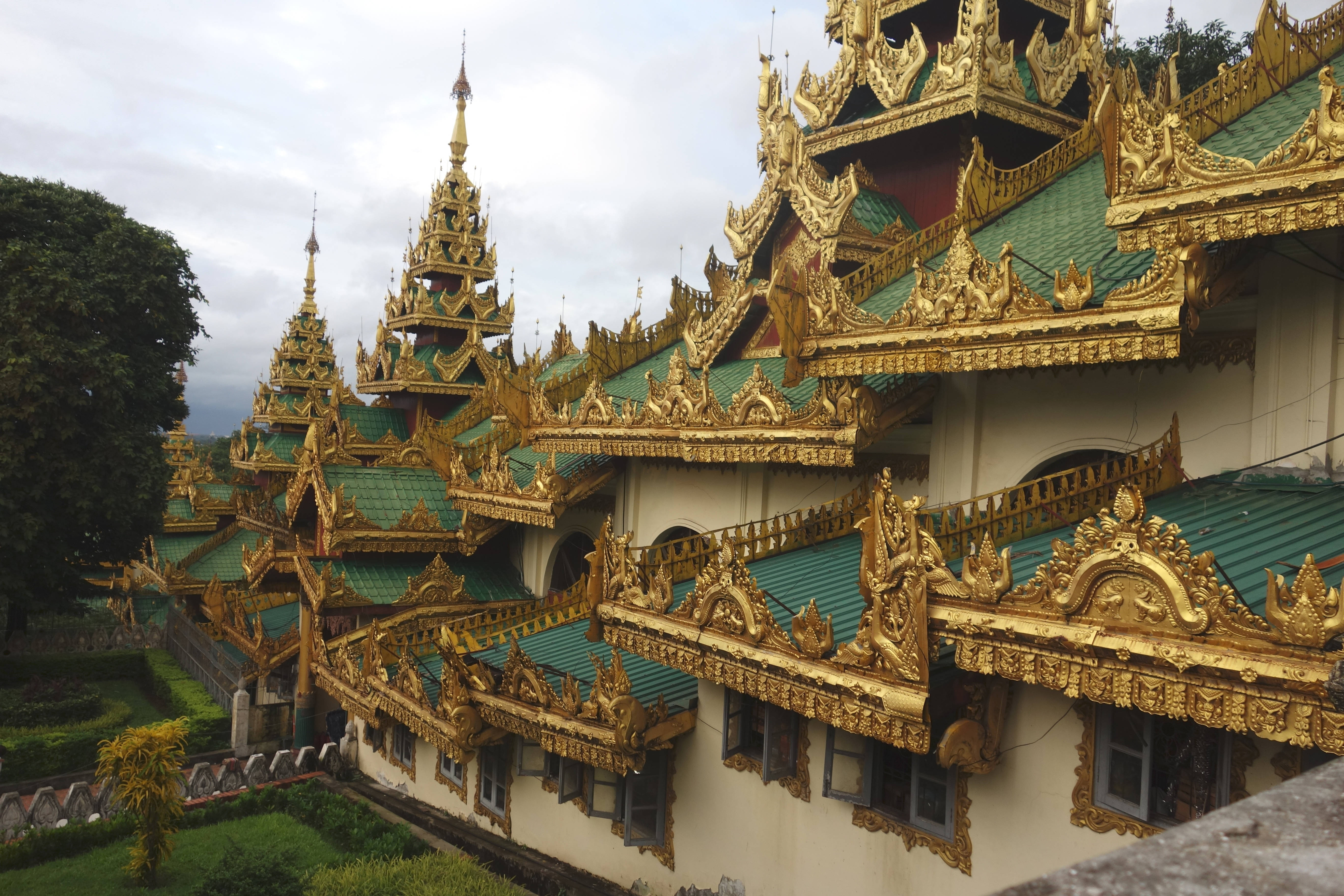 Yangon 桌布 壁紙 — 緬甸寺廟建築群的金色尖頂和華麗的綠色屋頂在陰天中聳立，下方是茂密的綠植。