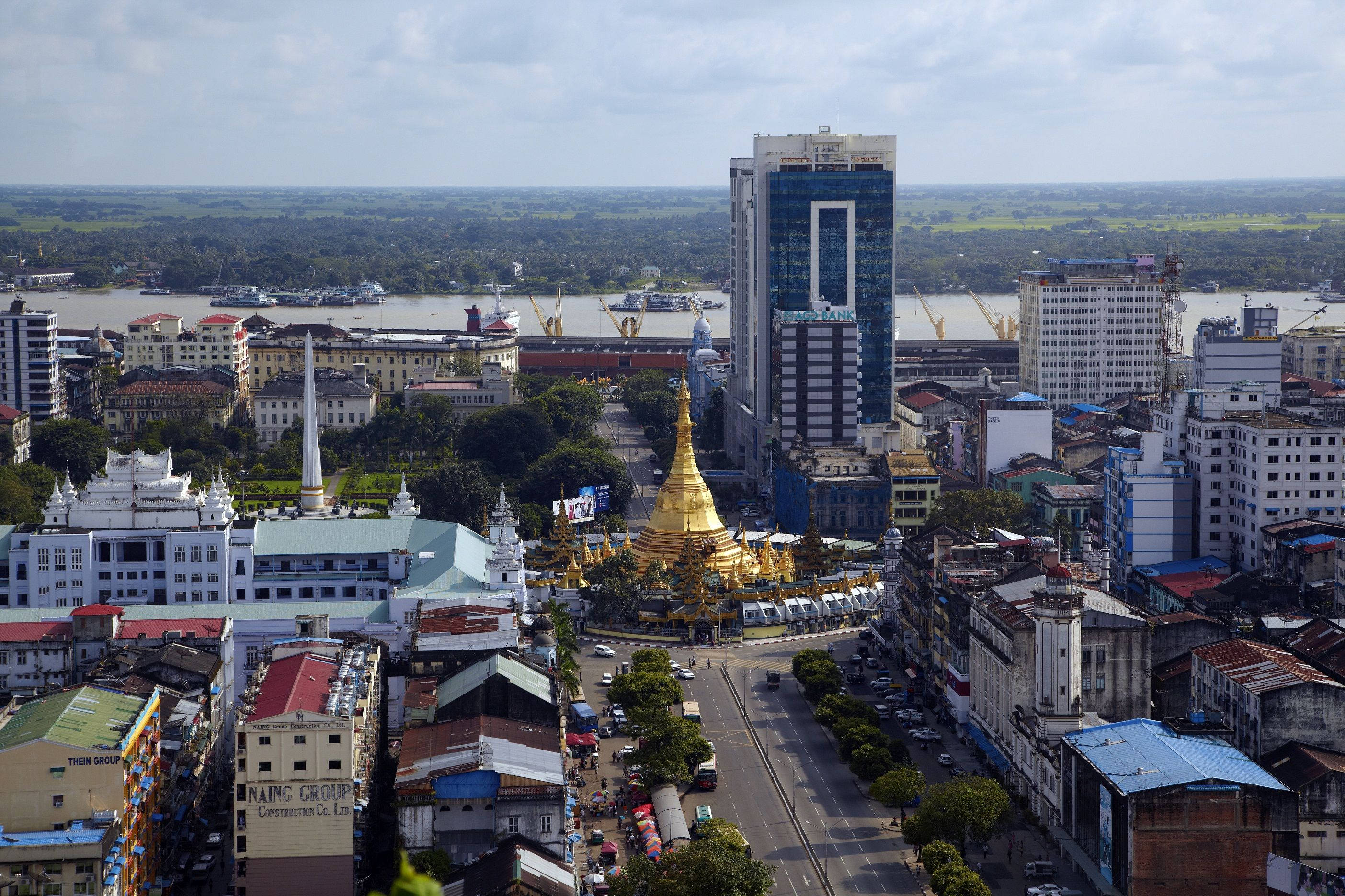 Yangon 桌布 壁紙 — 繁忙城市景觀的鳥瞰圖，一座金色寶塔在日光下閃耀，周圍是沿著寬闊河流的現代和傳統建築。