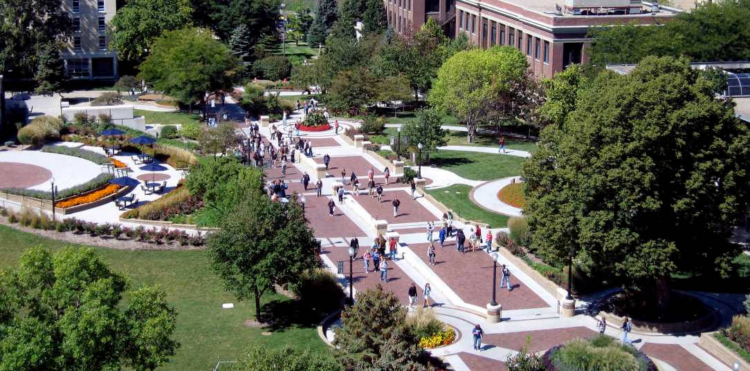 Nebraska 桌布 壁紙 — 繁忙大學校園的鳥瞰圖，學生沿著磚砌小徑行走，周圍環繞著茂密的綠樹和修剪整齊的花園。
