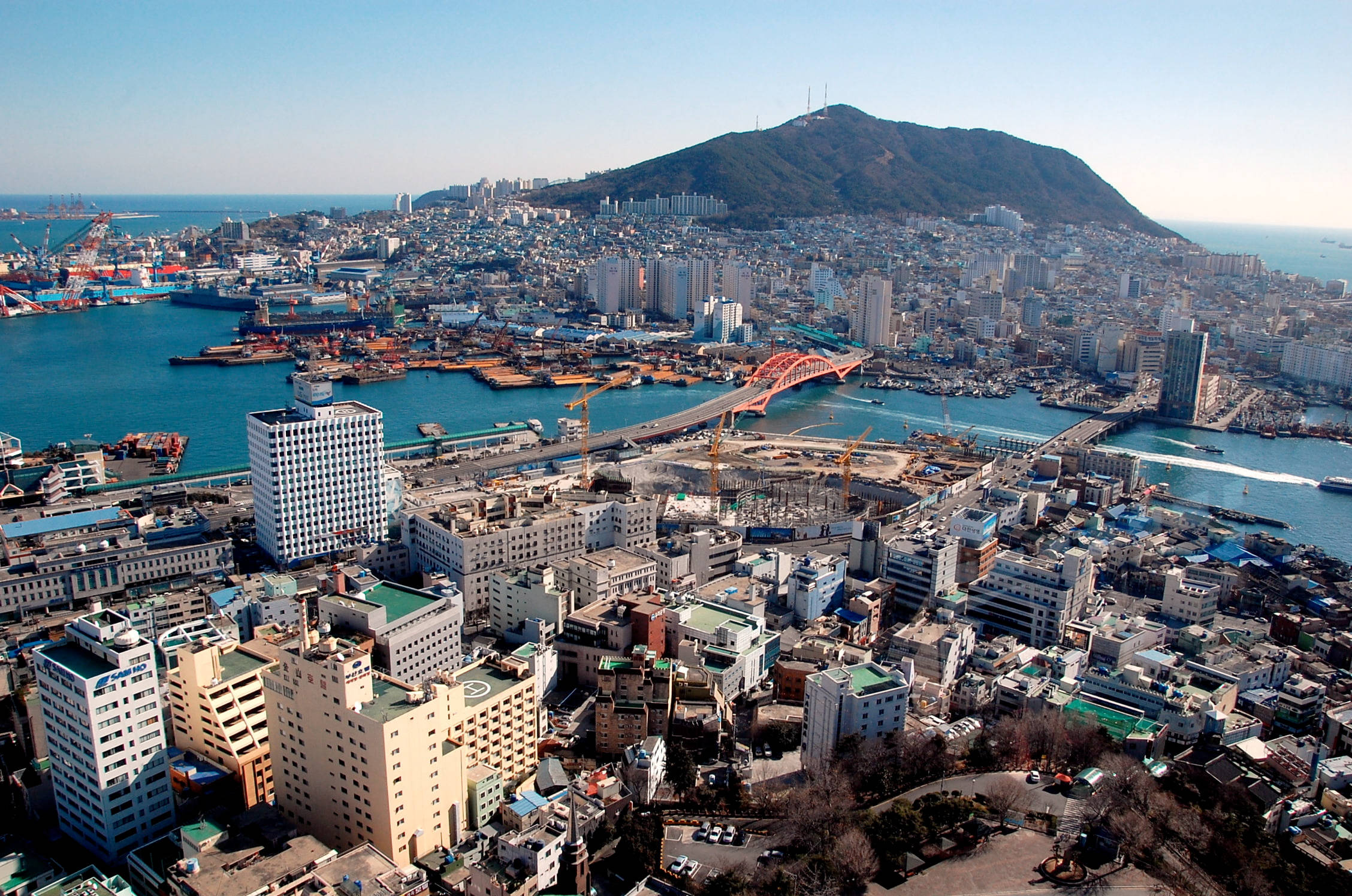 Busan 桌布 壁紙 — 繁忙沿海城市的鳥瞰圖，一座紅色橋樑橫跨碧綠海水，密集的城市建築後方升起一座綠色山丘。