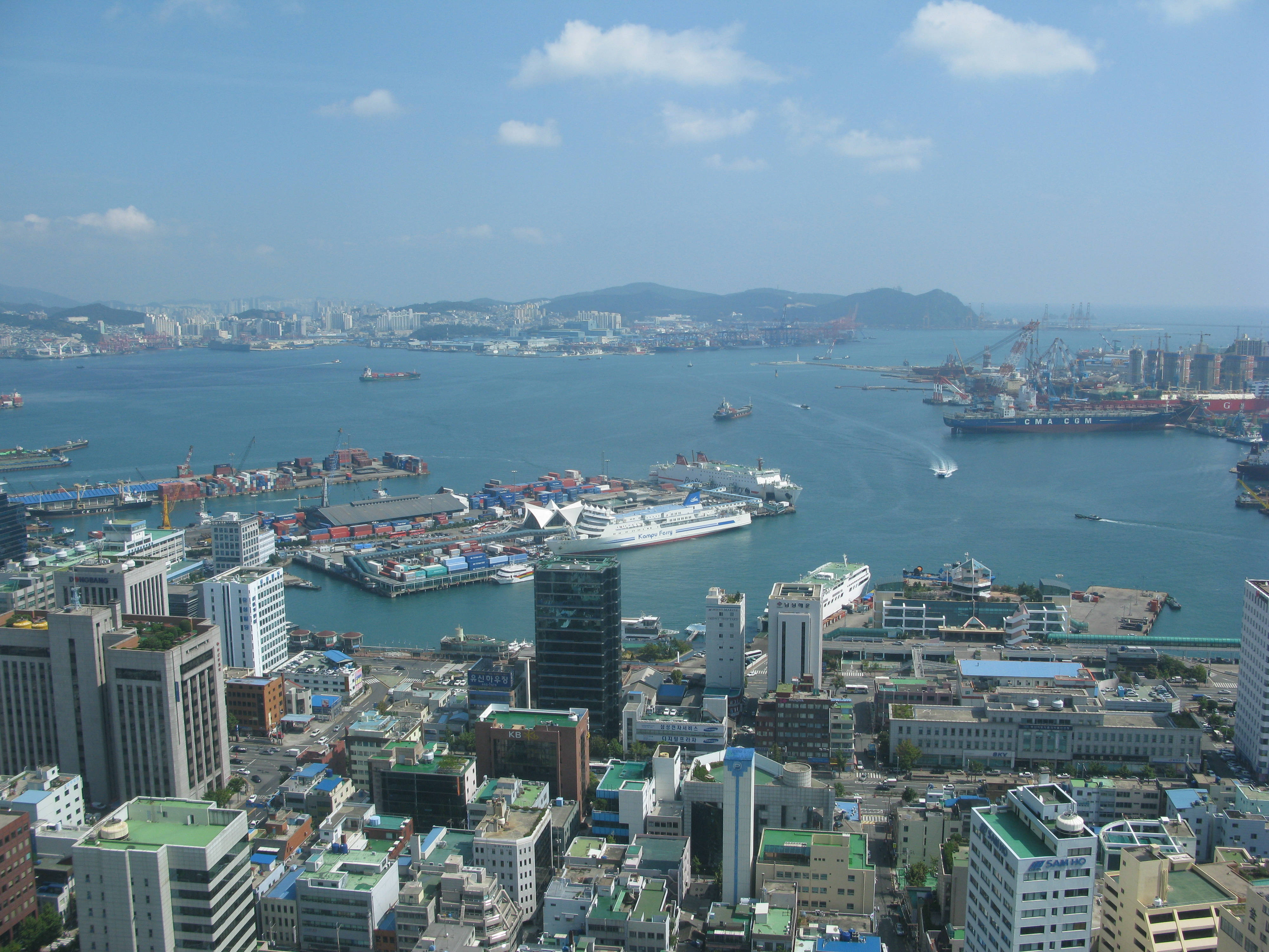 Busan 桌布 壁紙 — 繁忙港口城市的鳥瞰圖，擁有現代建築、貨船和清澈的藍天。