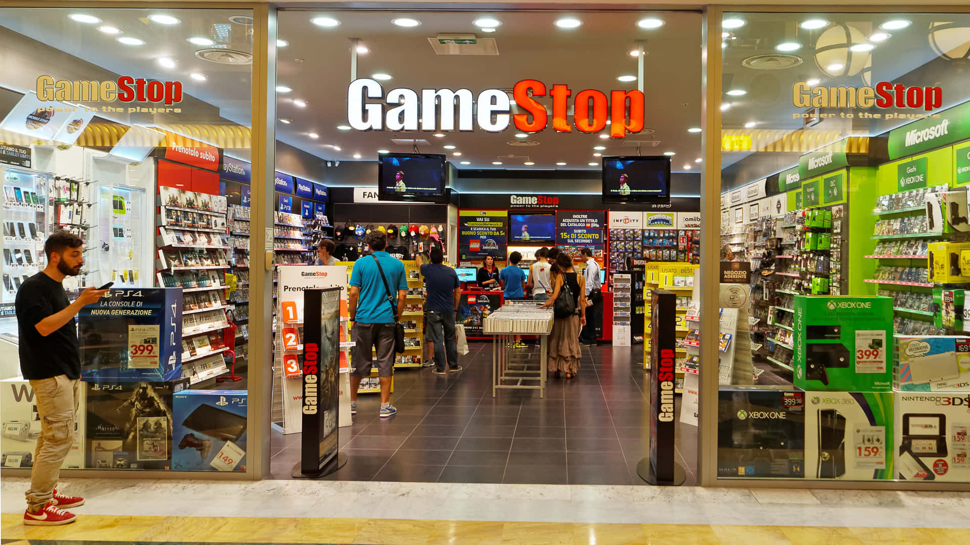 GameStop 桌布 壁紙 — 繁忙的GameStop商店入口，顧客在明亮的頂燈下瀏覽遊戲機和商品。