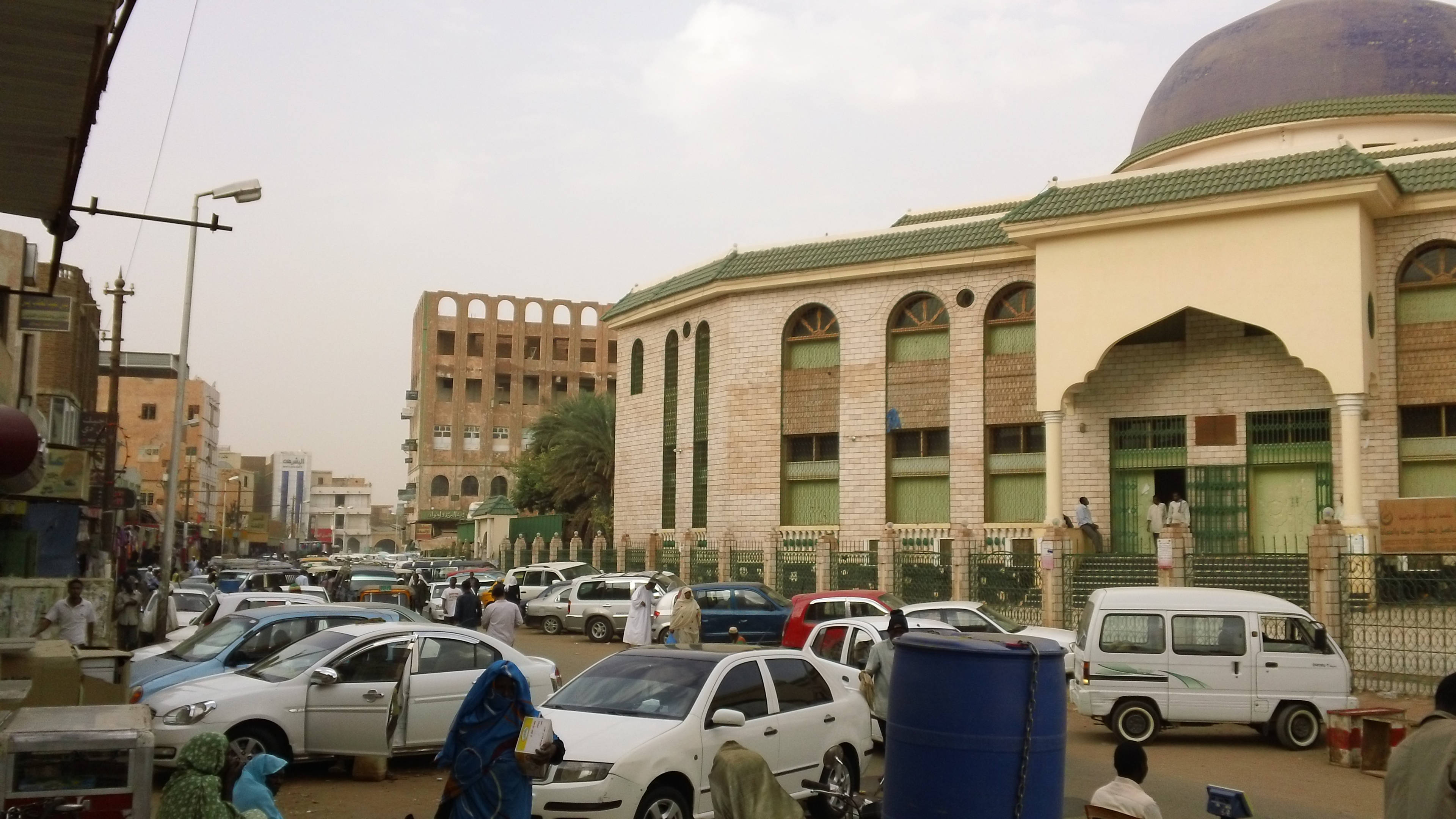 Sudan 桌布 壁紙 — 繁忙的城市街道場景，附近有一座藍色圓頂和綠色瓦頂的清真寺，停泊著汽車和行人。