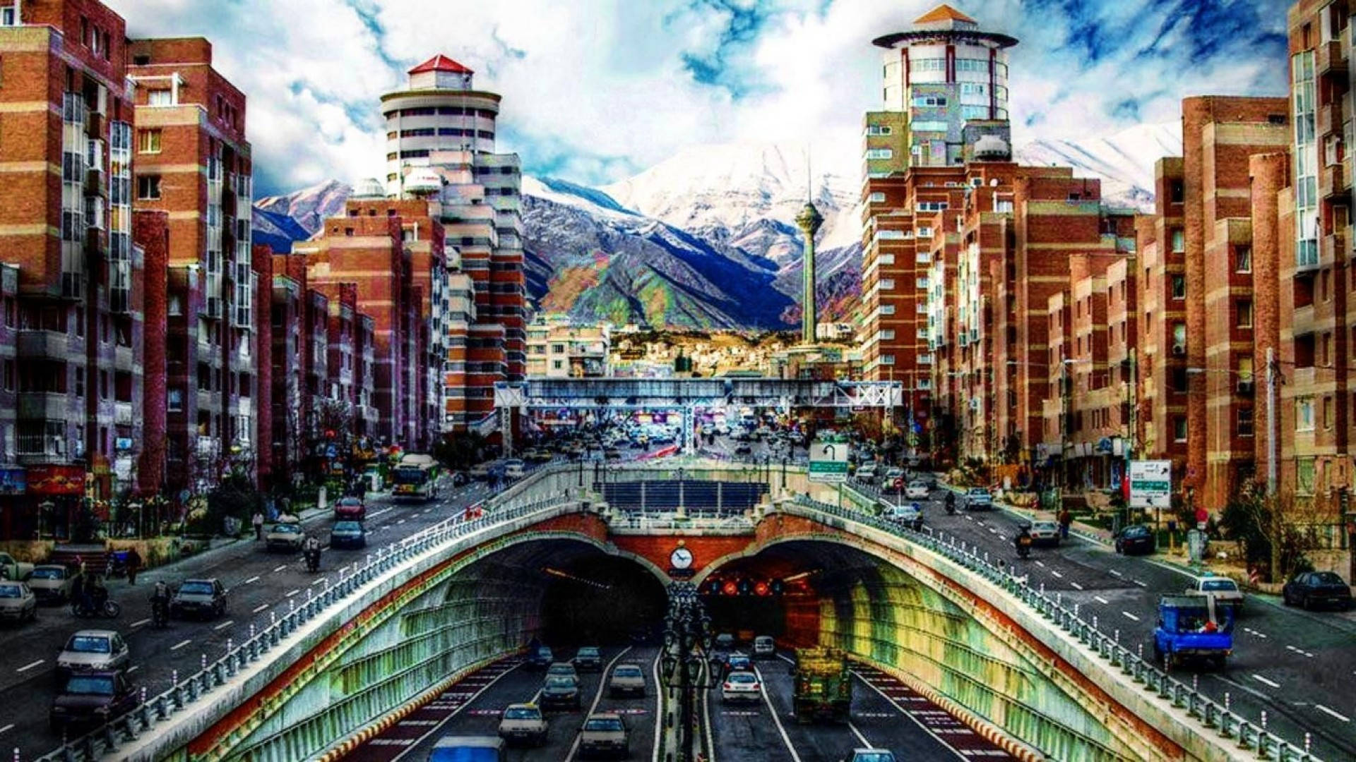 Tehran 桌布 壁紙 — 繁忙的城市高速公路隧道入口，由紅磚建築環繞，背後有積雪的山脈在陰天天空下聳立。