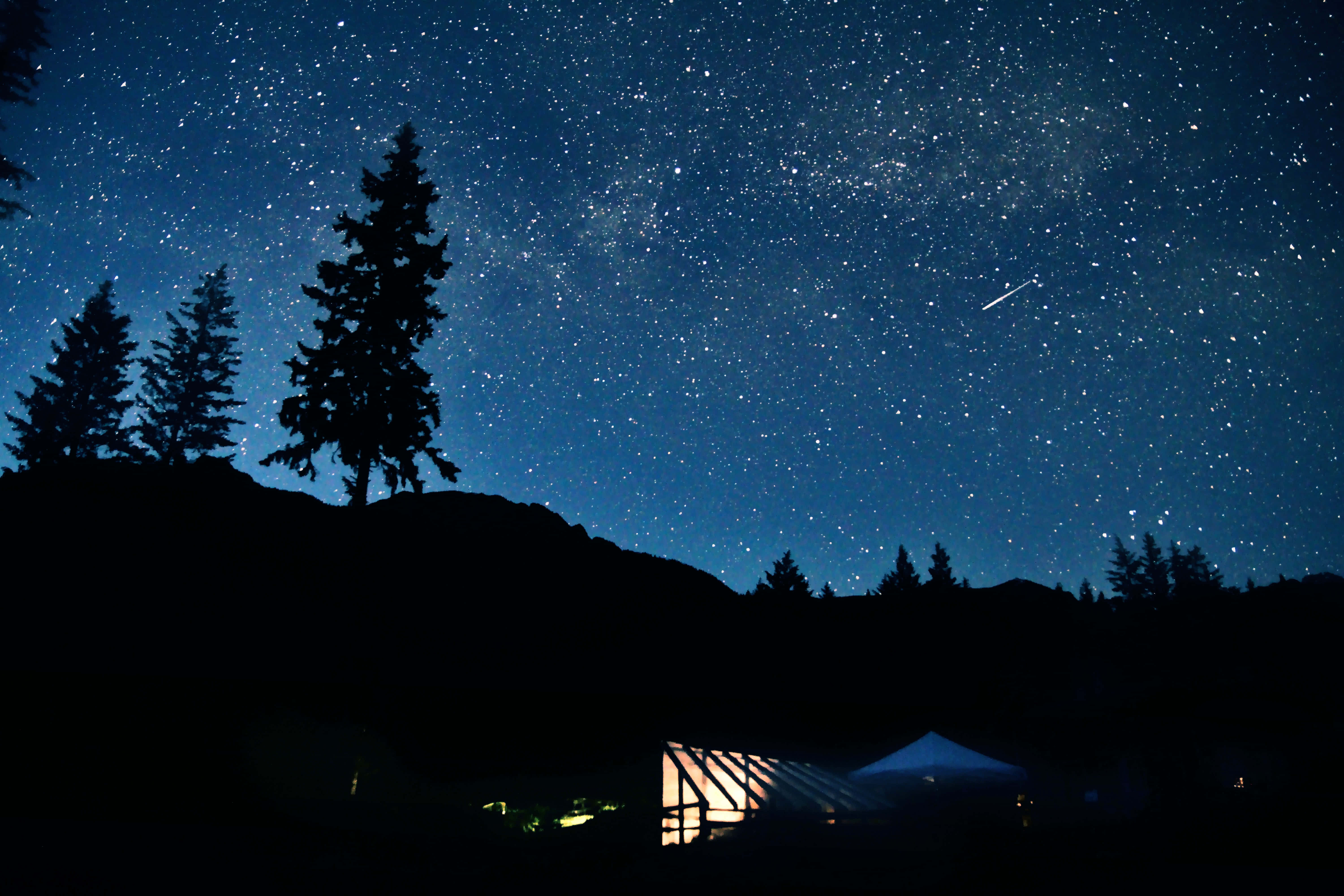 Silhouette 桌布 壁紙 — 繁星點點的夜空，一顆流星劃過剪影般的松樹和發光的露營帳篷上方。