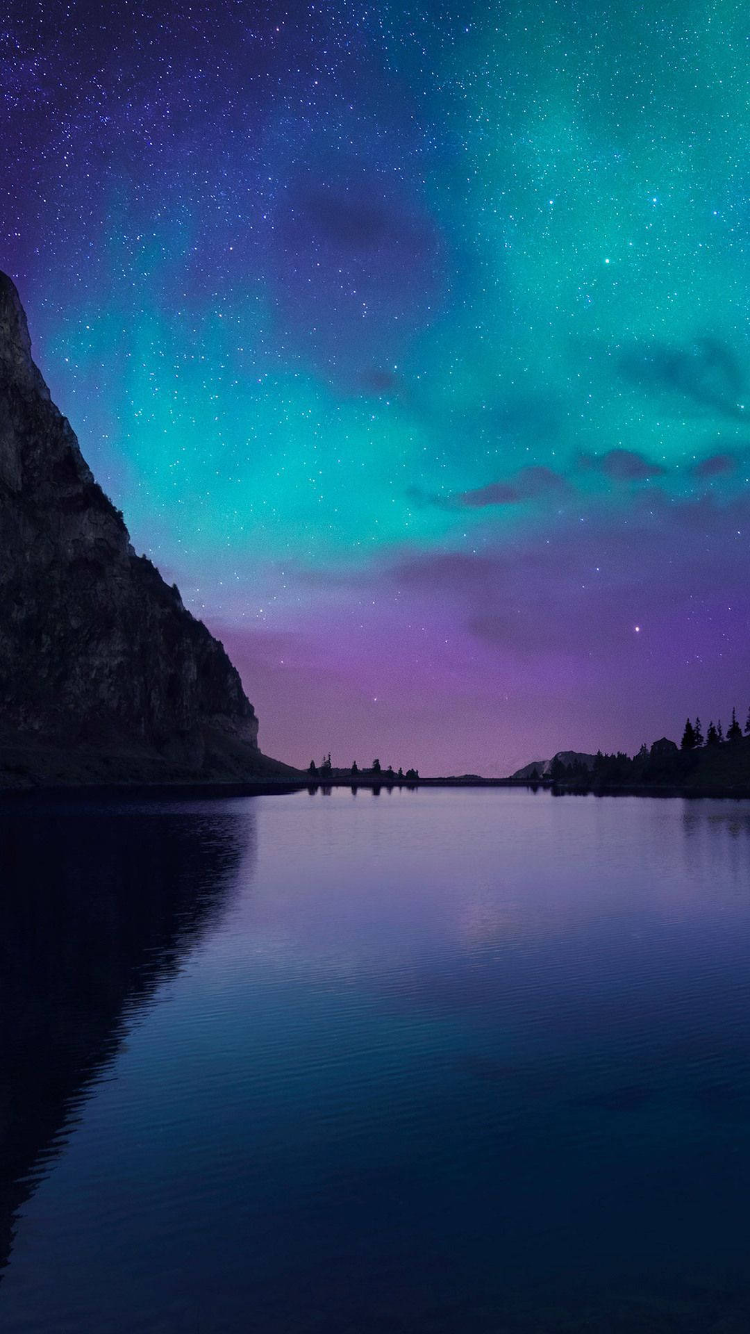 iOS 15 桌布 壁紙 — 繁星點點的夜空，充滿活力的綠色和紫色極光映照在平靜的湖面上，旁邊是黑暗的懸崖。