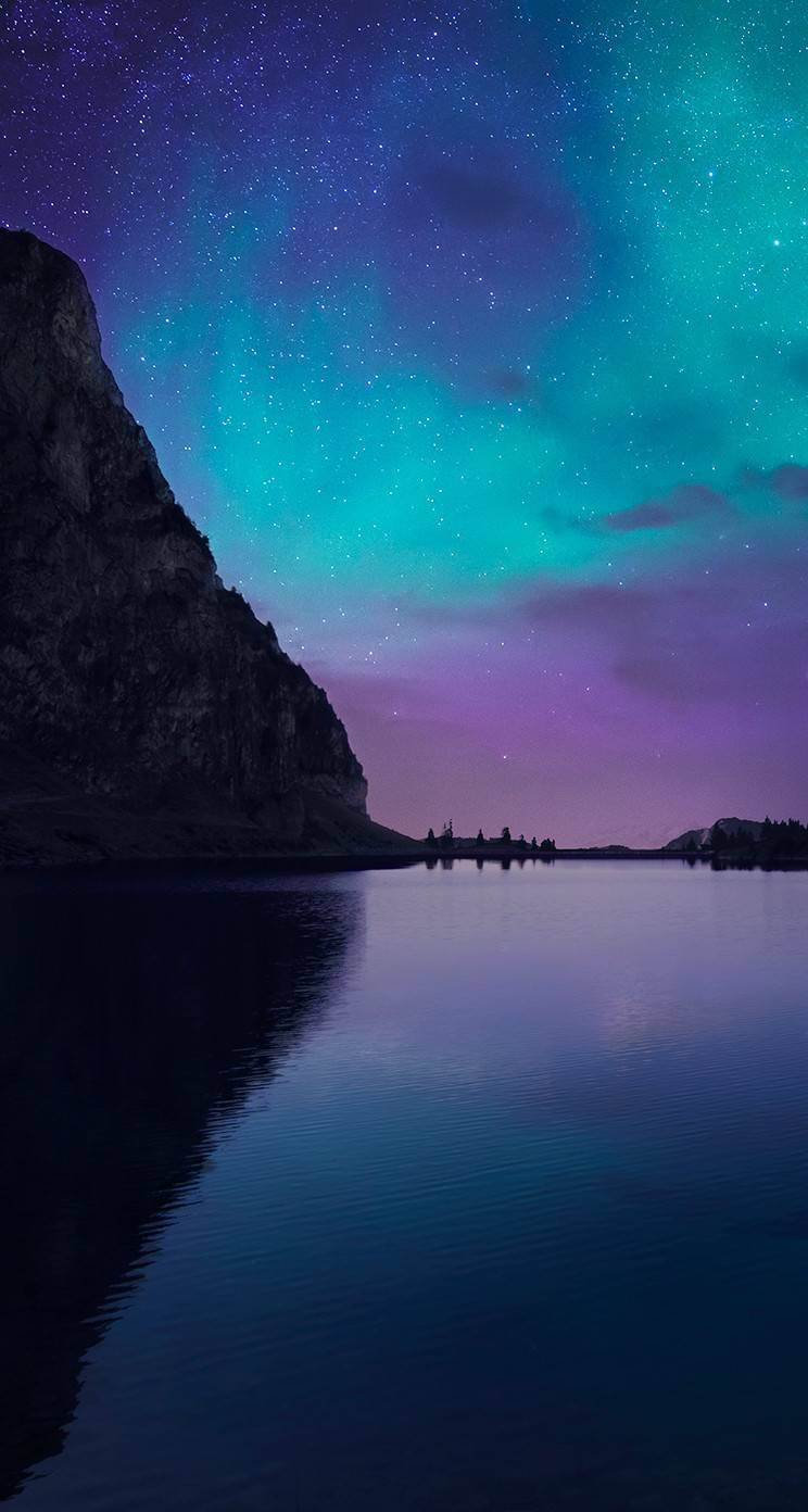 iOS 8 桌布 壁紙 — 繁星點點的夜空，充滿活力的綠色和紫色極光完美地映照在平靜的湖面上，旁邊是黑暗的岩石懸崖。