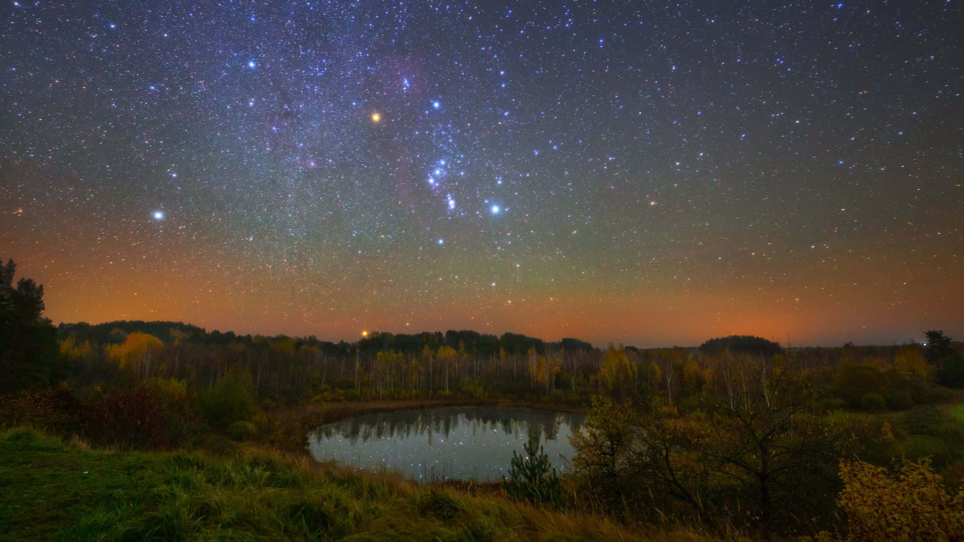 星空動畫 桌布 壁紙 — 繁星點點的夜空，帶有充滿活力的橙色極光，籠罩在一個被秋樹環繞的寧靜池塘上。