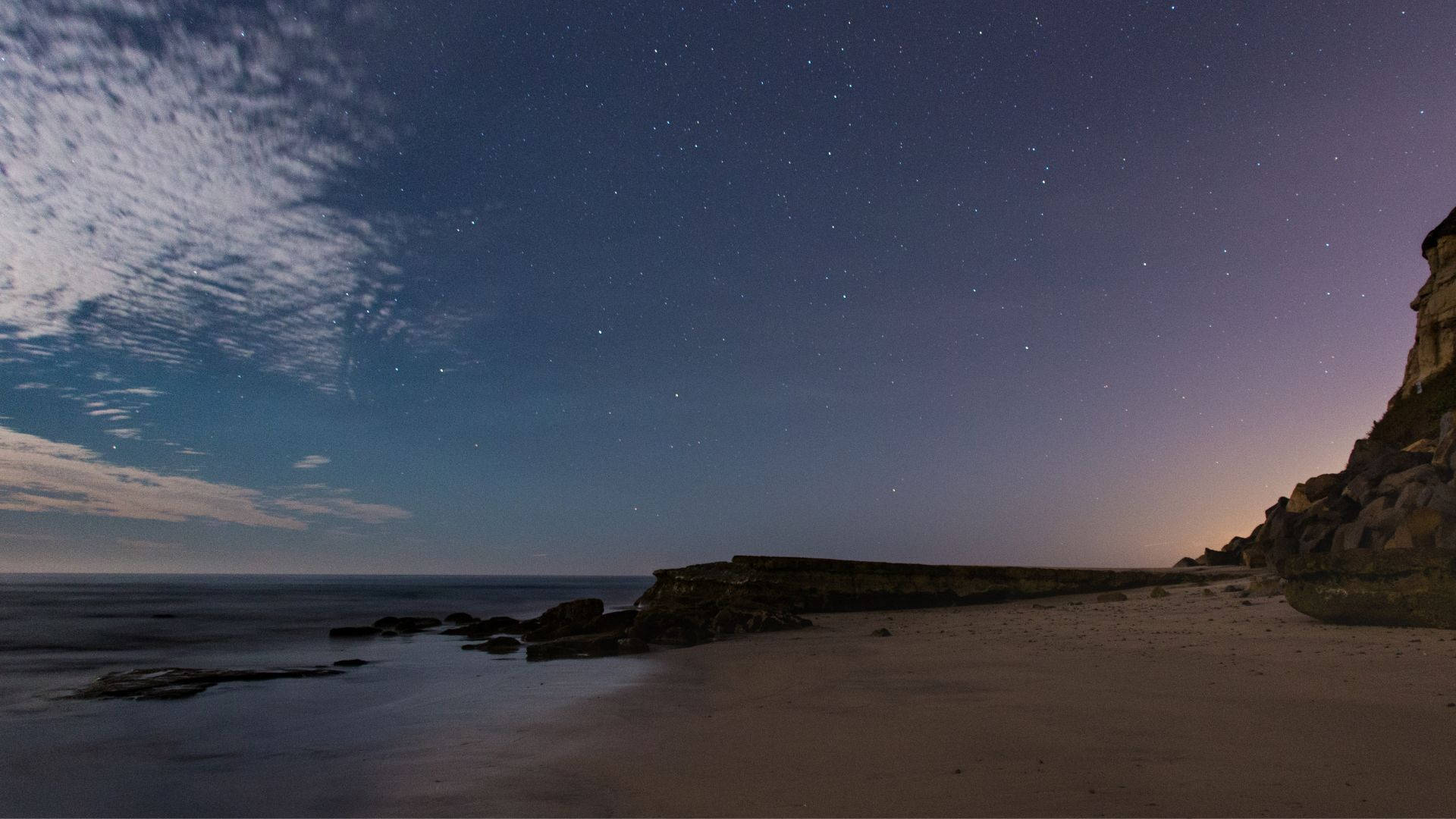 電腦 桌布 壁紙 — 繁星點點的夜空，薄雲飄浮，籠罩著寧靜的海灘和海洋旁的岩石峭壁。