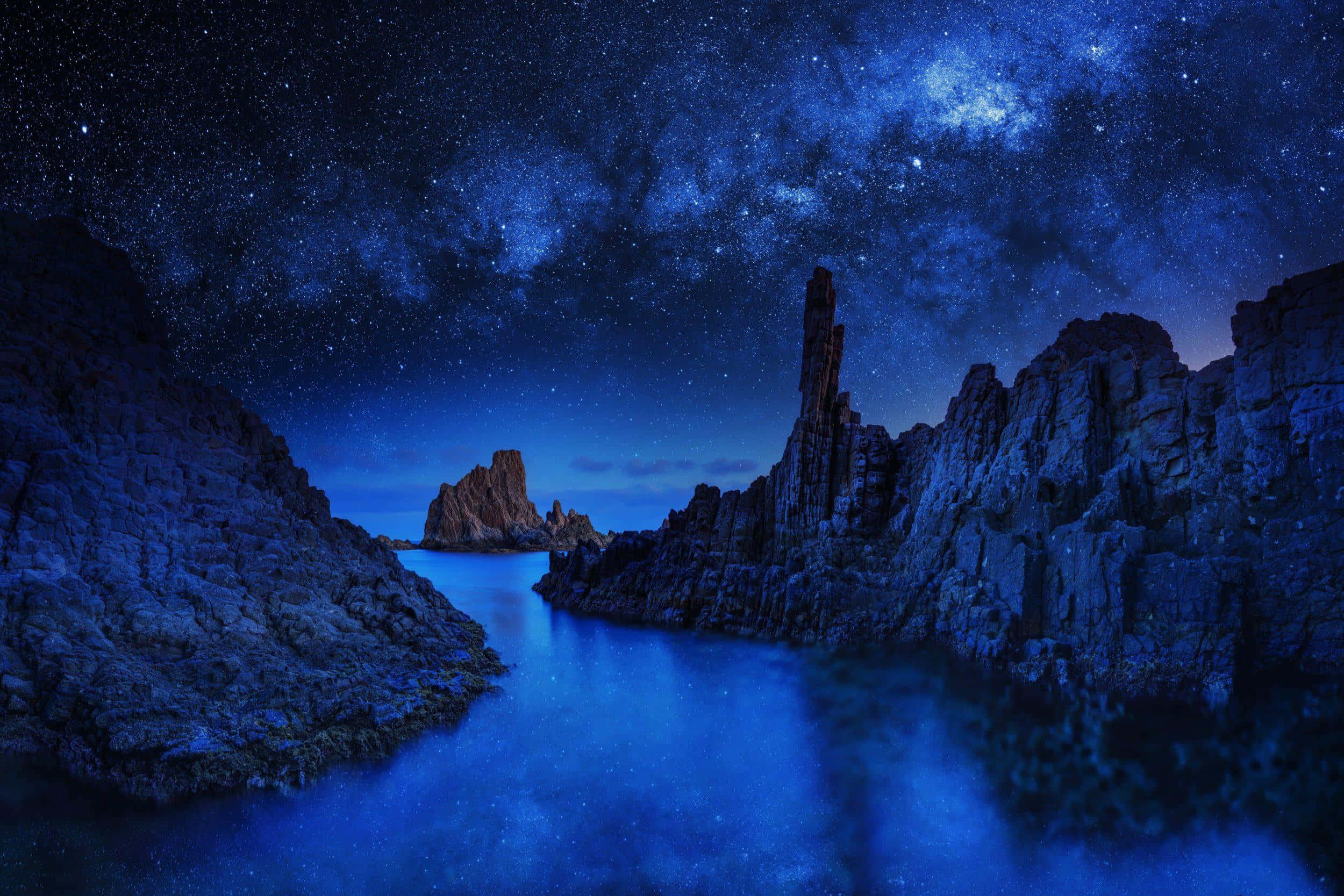 夜空 桌布 壁紙 — 繁星點點的夜空，銀河在平靜的藍色海洋上空拱起，兩側是崎嶇的懸崖。