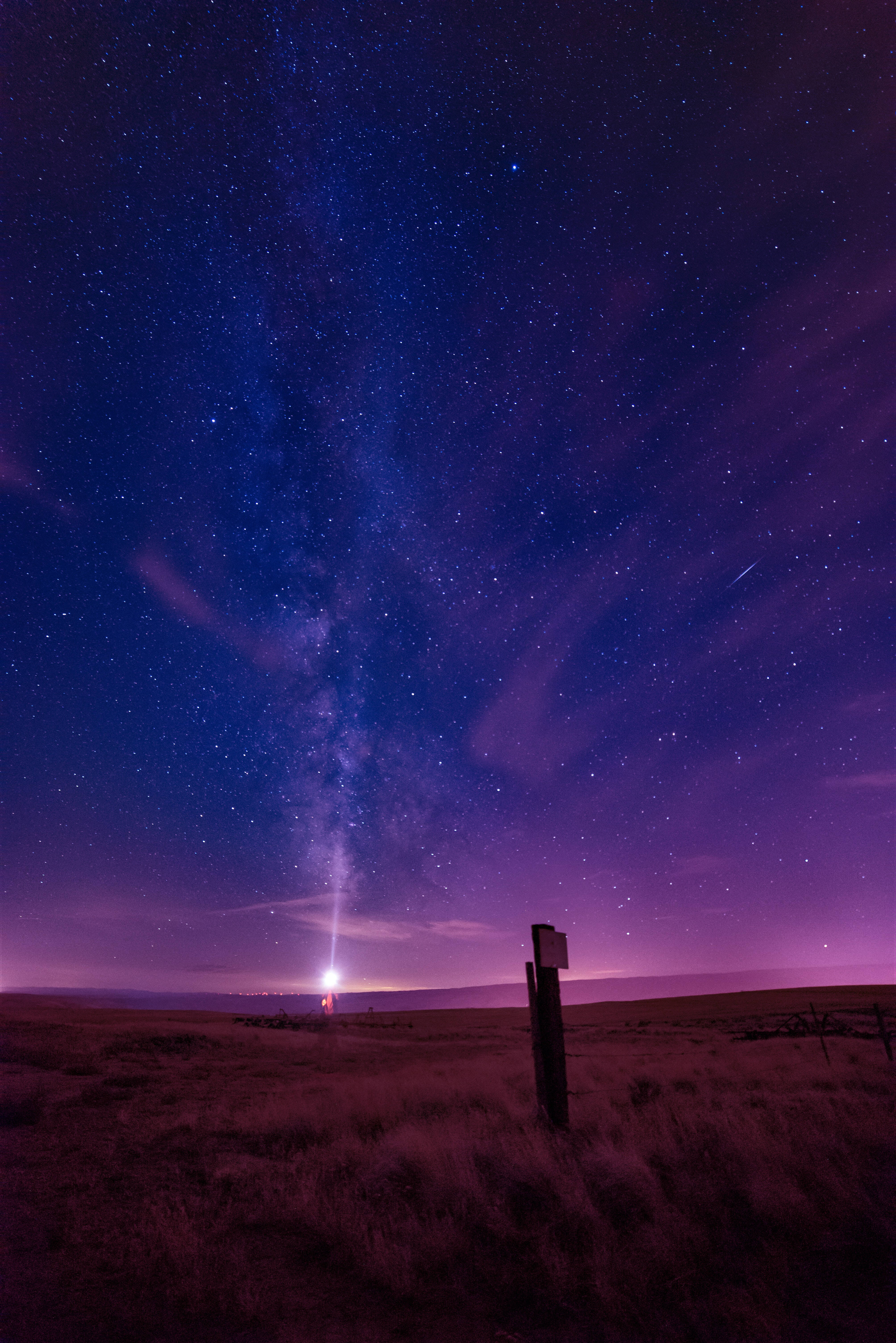 Pretty Galaxy 桌布 壁紙 — 繁星點點的夜空，銀河在深紫色沙漠景觀上空拱起，孤獨的籬笆柱和遠處的燈光。