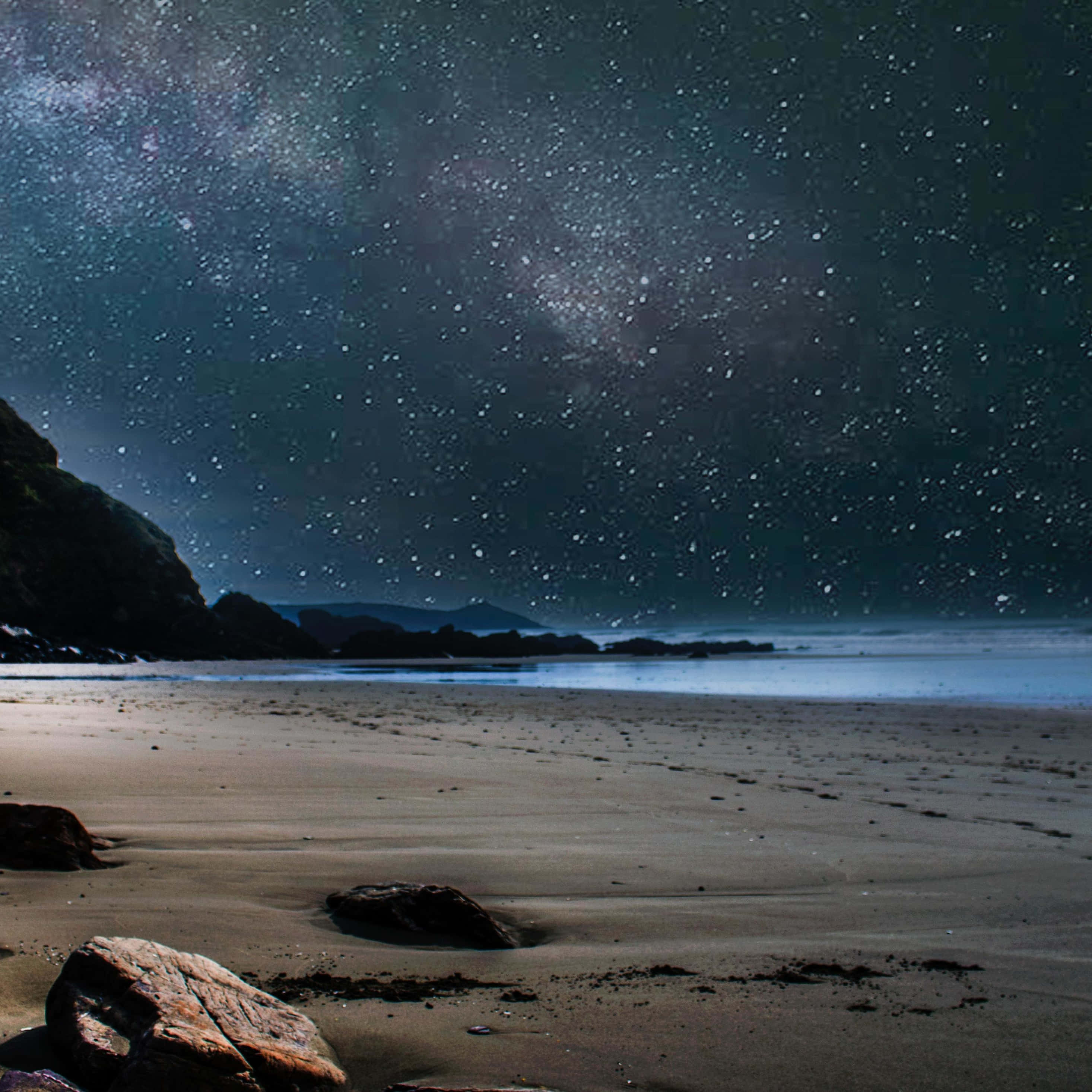 桌布 壁紙 — 繁星點點的夜空，銀河拱橋般橫跨在一個寧靜的海灘上，海灘上有深色的岩石和平靜的海水。