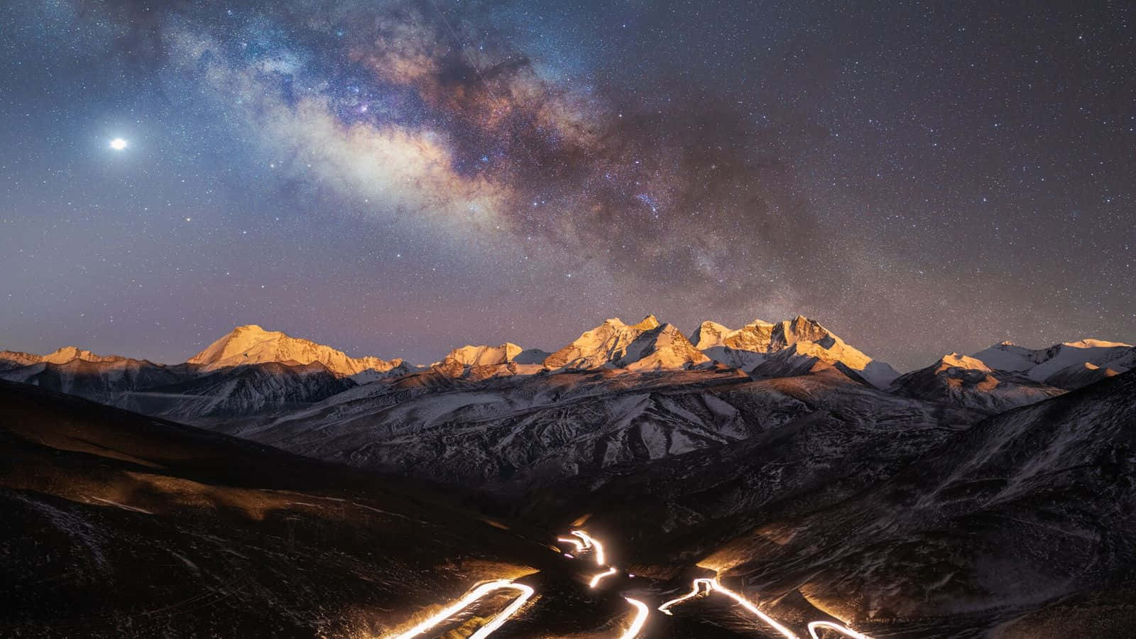 astronomy 桌布 壁紙 — 繁星點點的夜空，銀河拱跨在月光和下方發光道路痕跡照亮的積雪山峰之上。