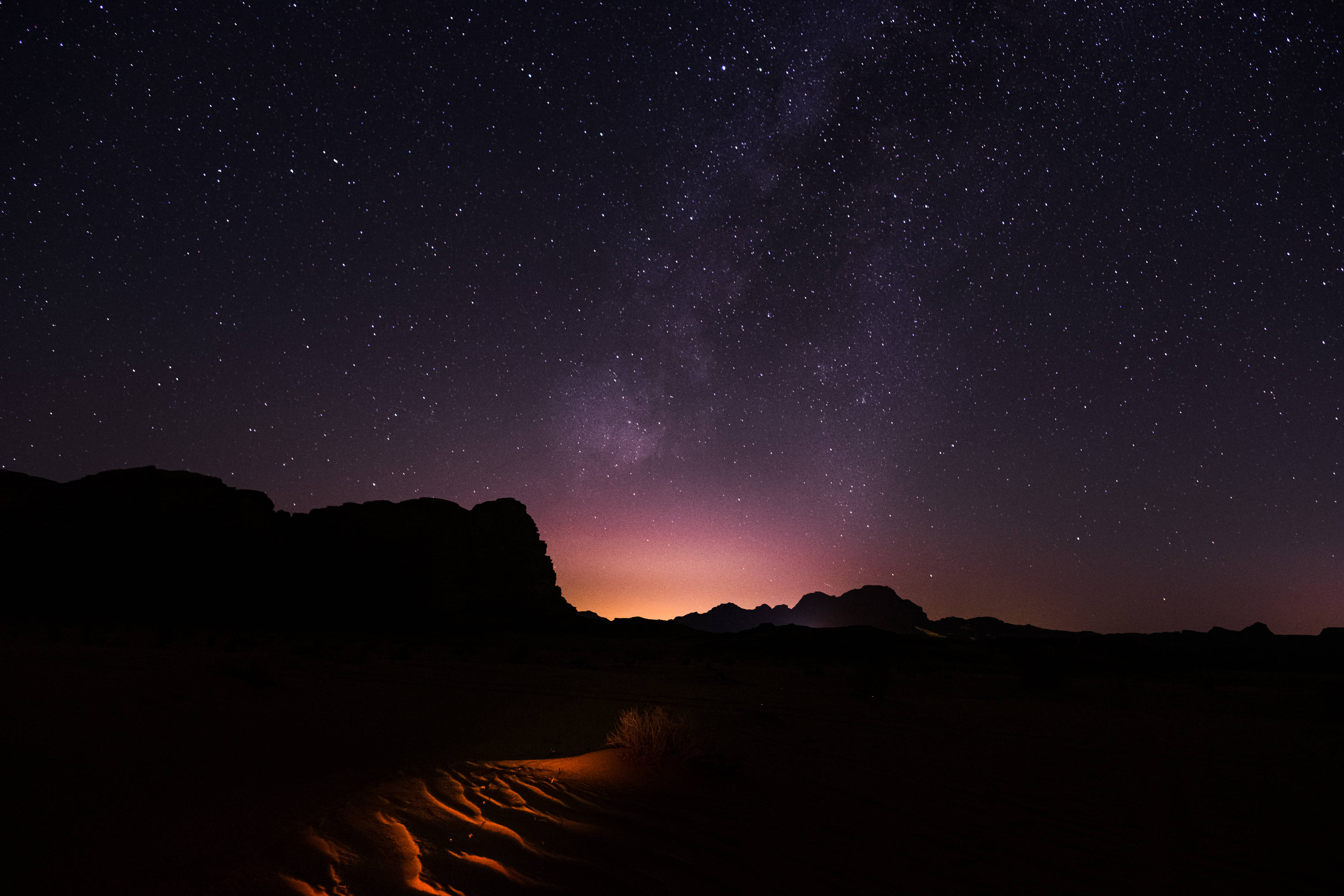 Jordan 桌布 壁紙 — 繁星點點的夜空，銀河拱跨黑暗的沙漠岩層和發光的橙色沙丘。