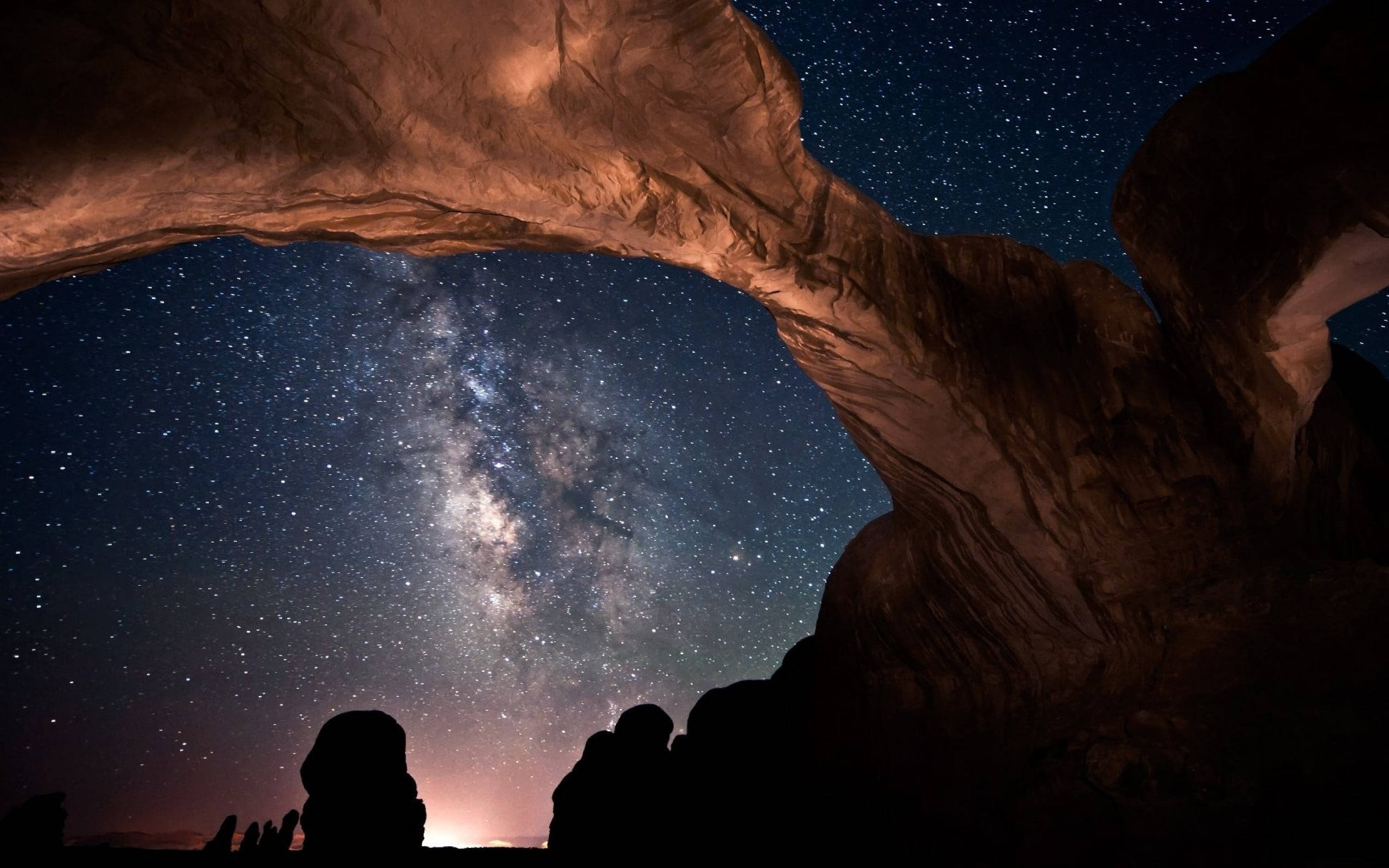銀河 桌布 壁紙 — 繁星點點的夜空，銀河穿過巨大的砂岩地貌拱起，剪影人物仰望。
