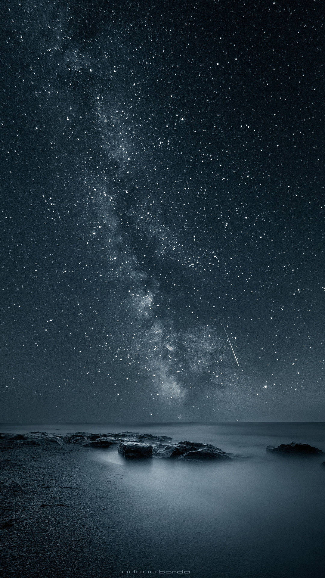 iPhone 8 live 桌布 壁紙 — 繁星點點的夜空，銀河與流星在平靜的海洋和前景的深色岩石上方拱起。
