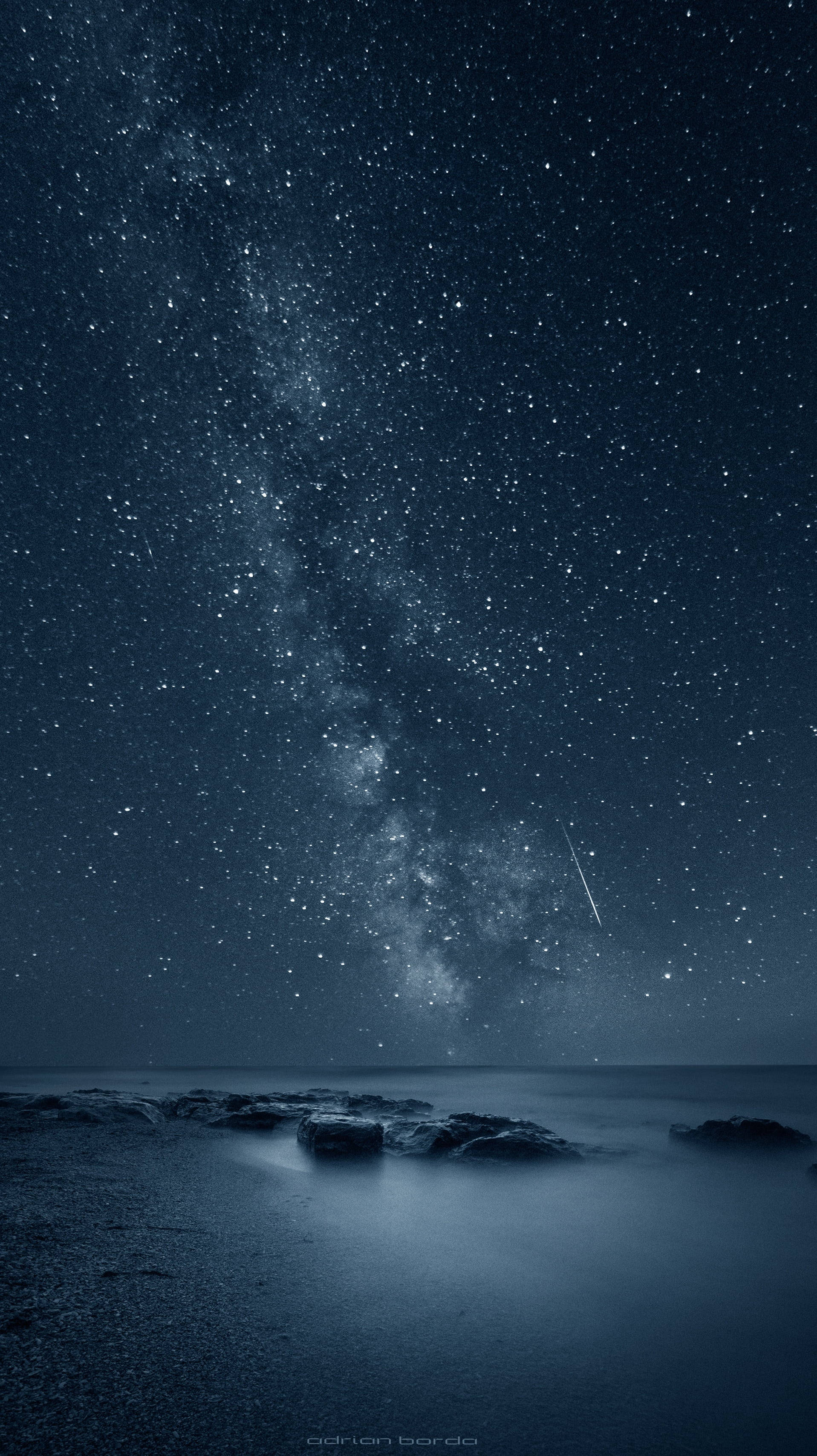 銀河 iPhone 桌布 壁紙 — 繁星點點的夜空，銀河與流星在平靜的海洋和前景的黑色岩石上拱起。