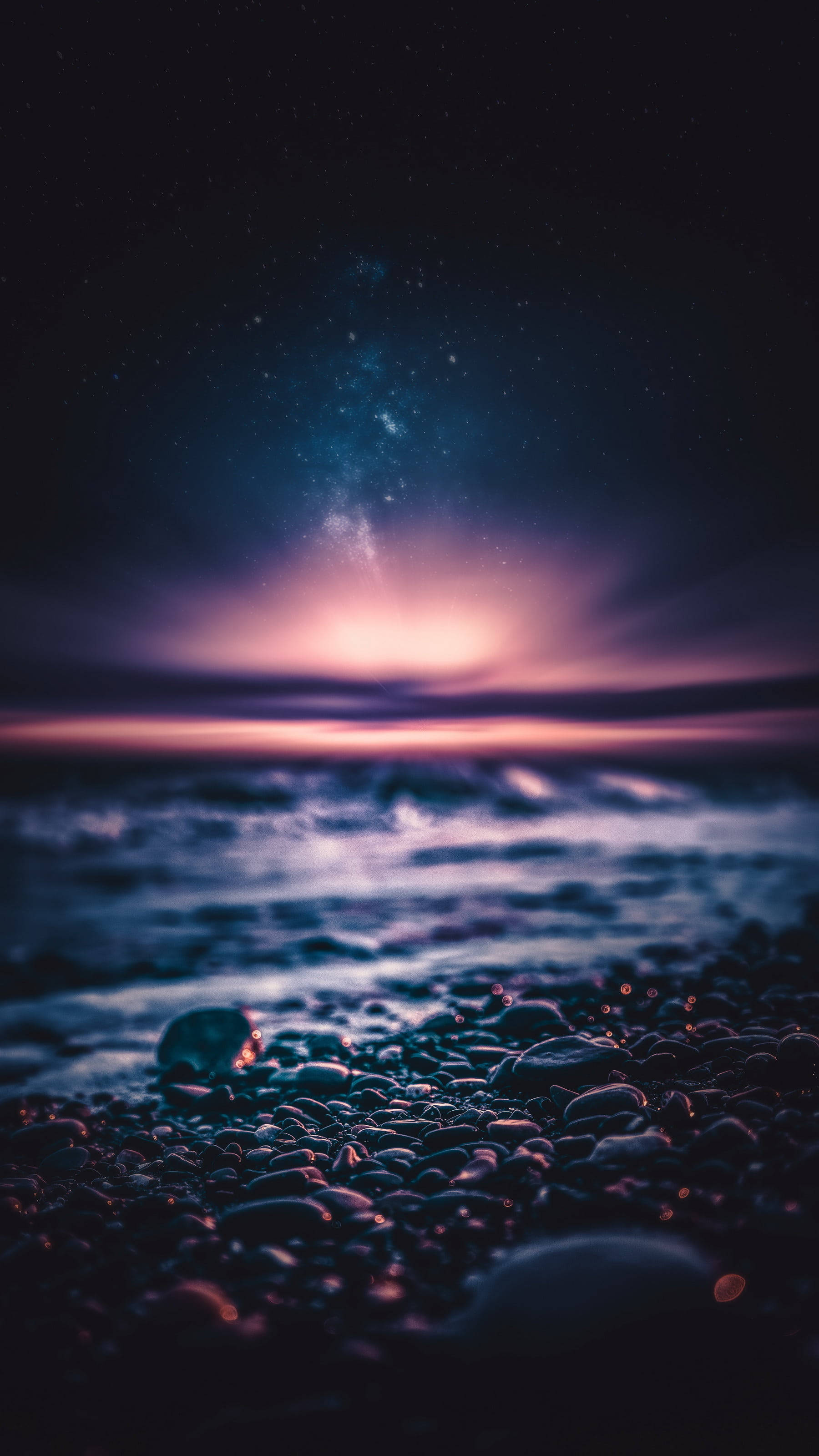 桌布 壁紙 — 繁星點點的夜空，閃爍的銀河拱跨在深色鵝卵石海灘上，海浪輕輕拍打岸邊。