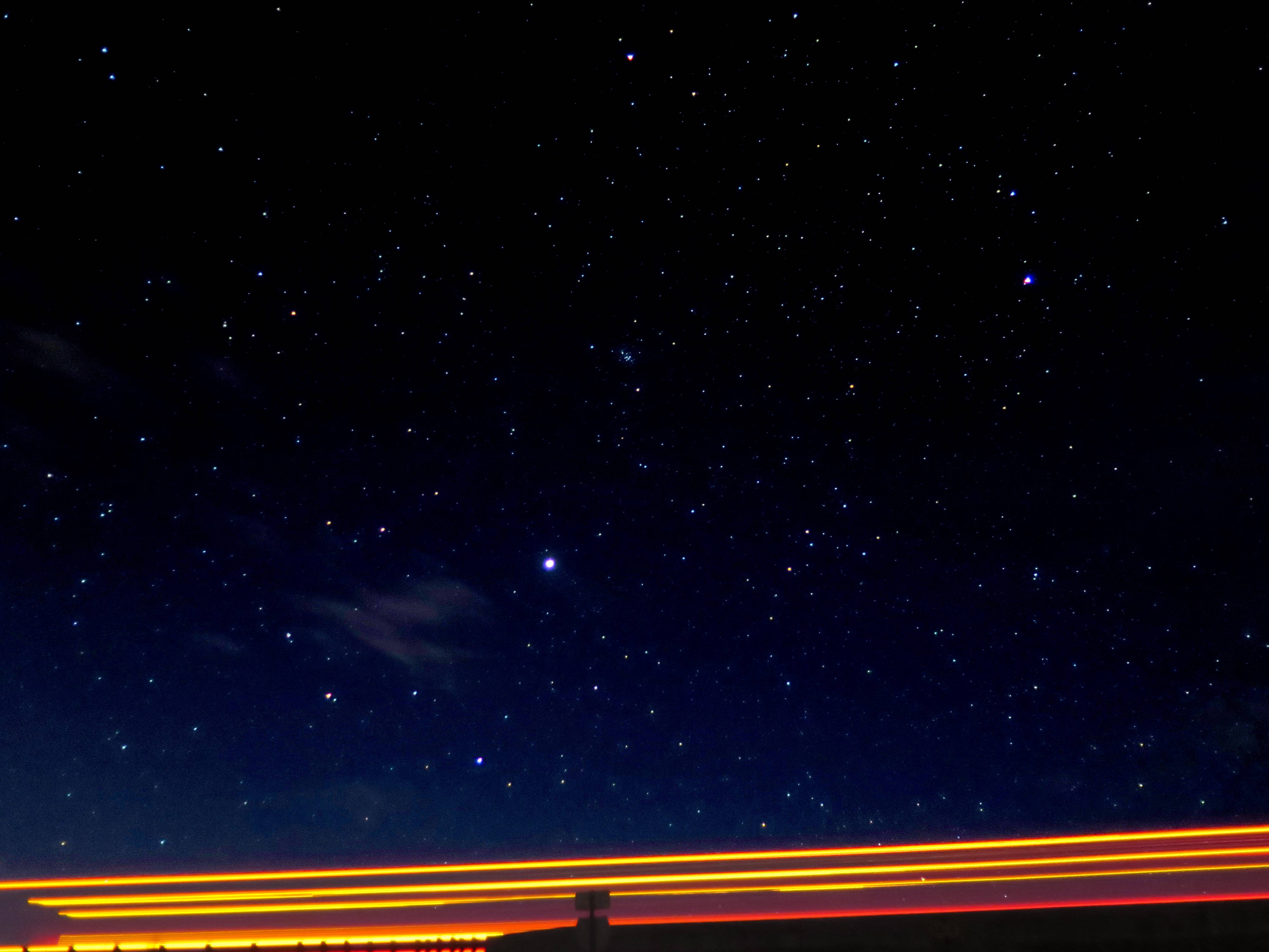 夜空 桌布 壁紙 — 繁星點點的夜空，黑暗地平線下方有車輛經過時留下的橙色發光光跡。