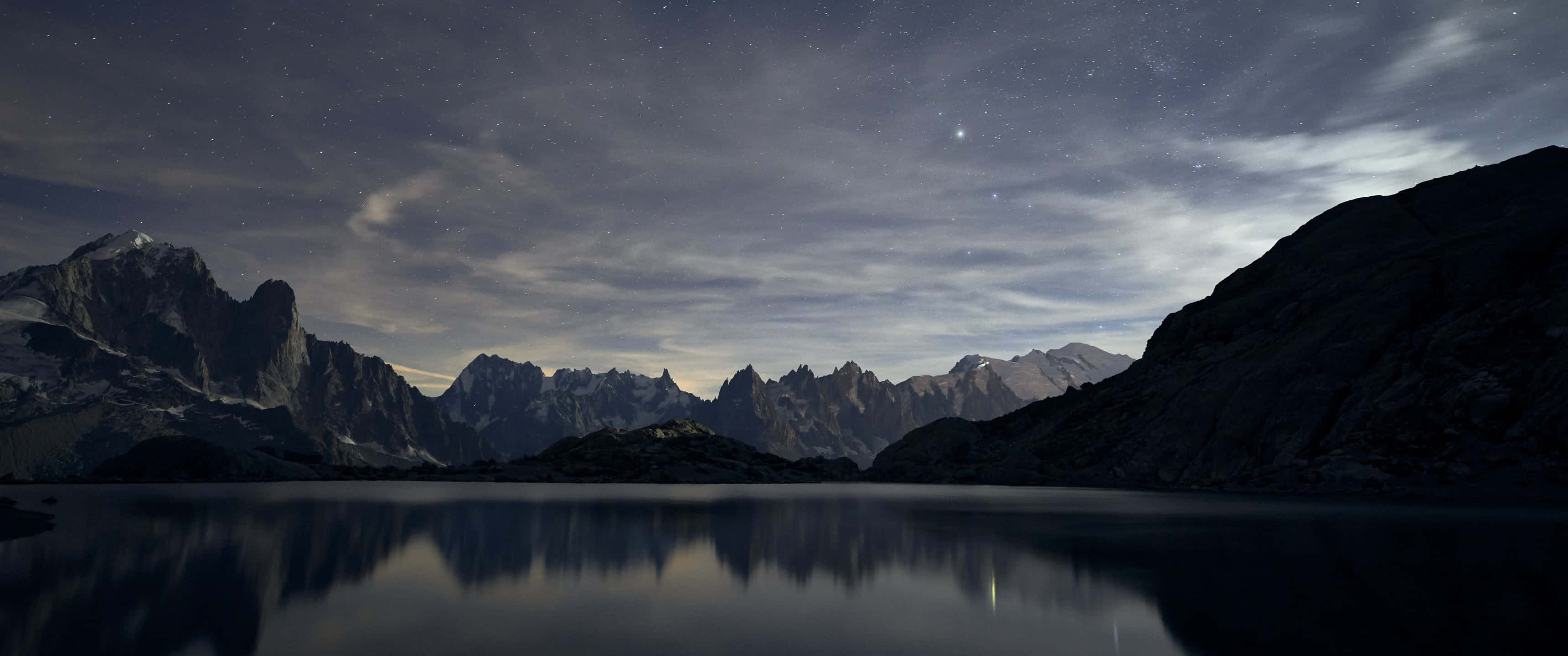 3440x1440p Oled 桌布 壁紙 — 繁星點點的夜空倒映在平靜的湖面上，周圍是鋸齒狀的山峰，籠罩在柔和的暮色雲層下。