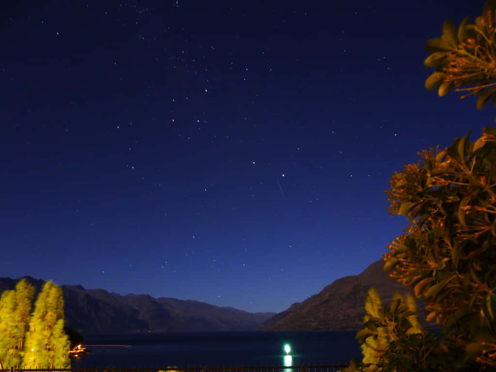 風景 桌布 壁紙 — 繁星點點的夜空在寧靜的湖泊上方閃耀，周圍是發光的樹木和遠山。
