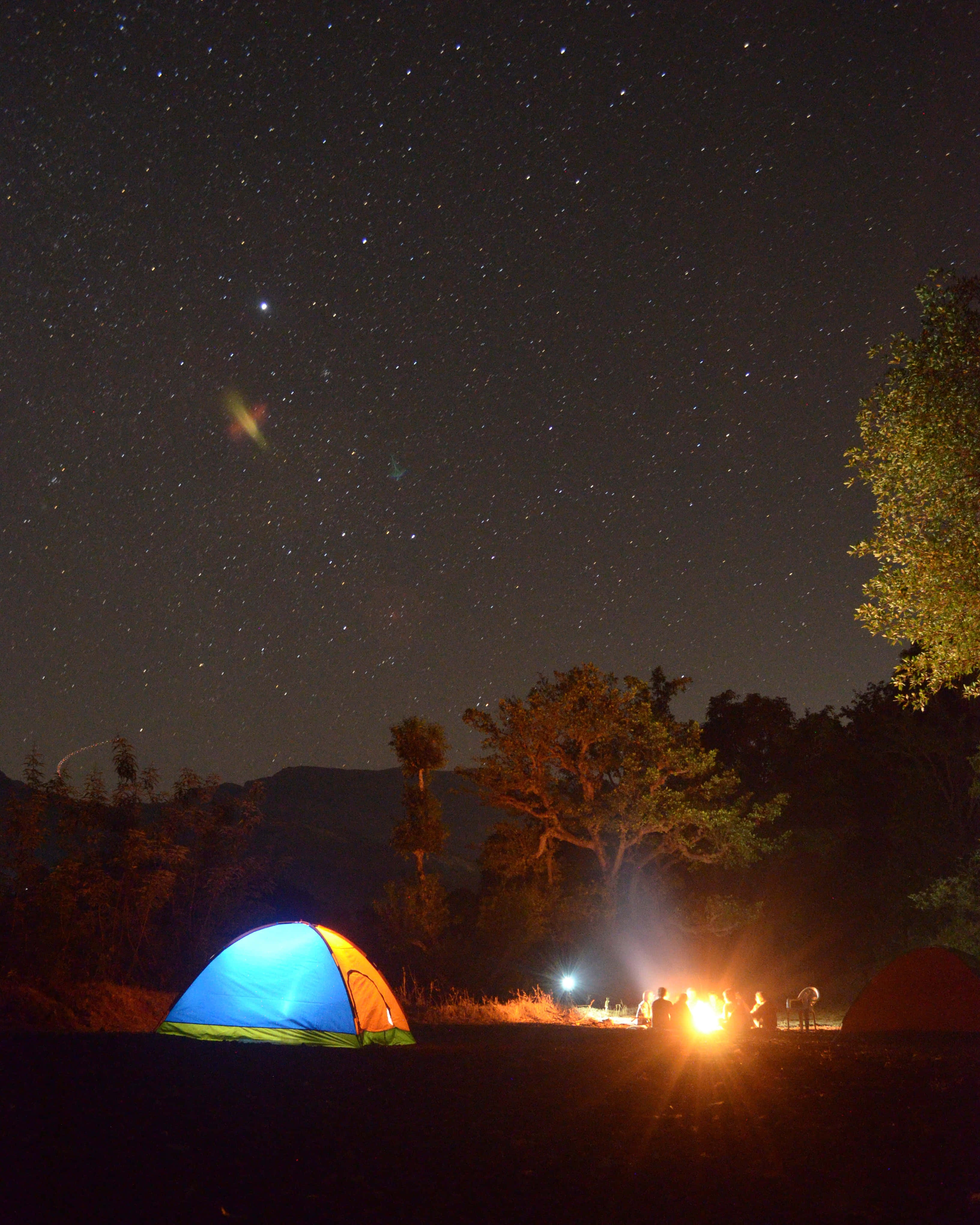 Bonfire 桌布 壁紙 — 繁星點點的夜空在營地上方閃耀，營地裡有一個發光的帳篷，人們圍坐在溫暖的營火旁。