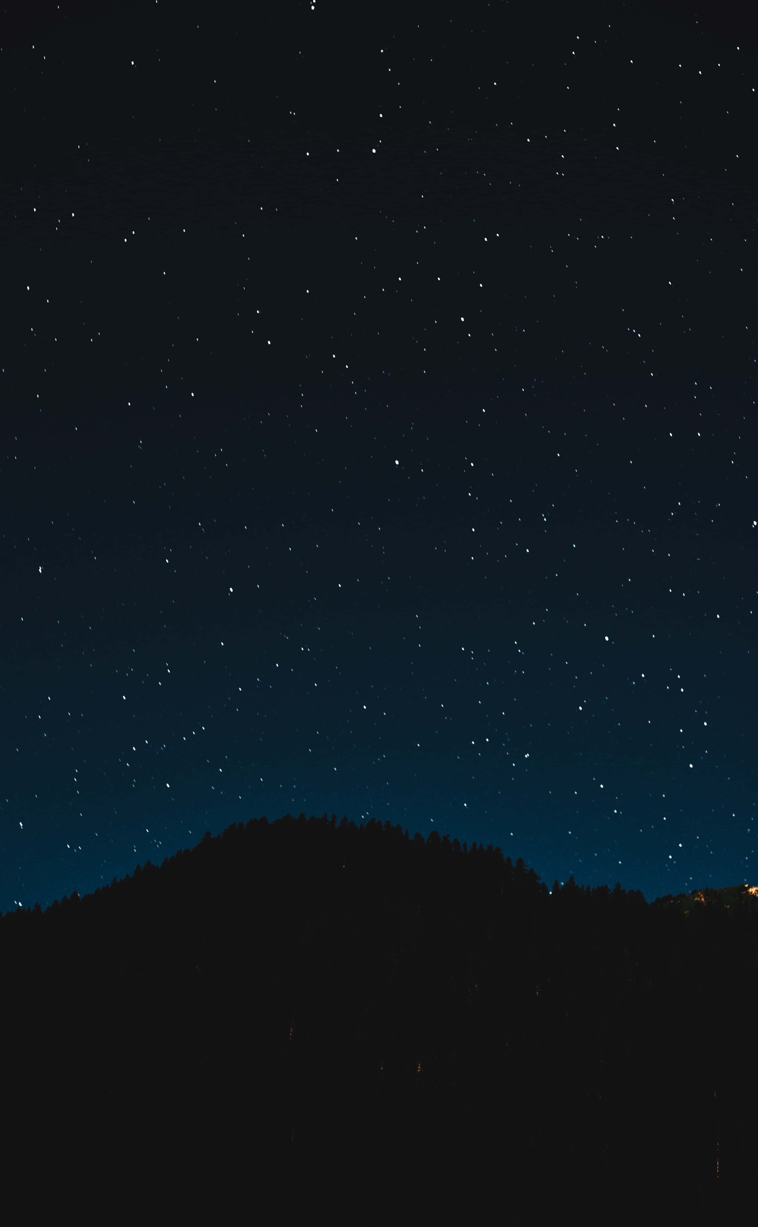 Starry Sky 桌布 壁紙 — 繁星點點的夜空在黑暗的森林山丘上方閃耀，遠方的燈光在地平線上閃爍。