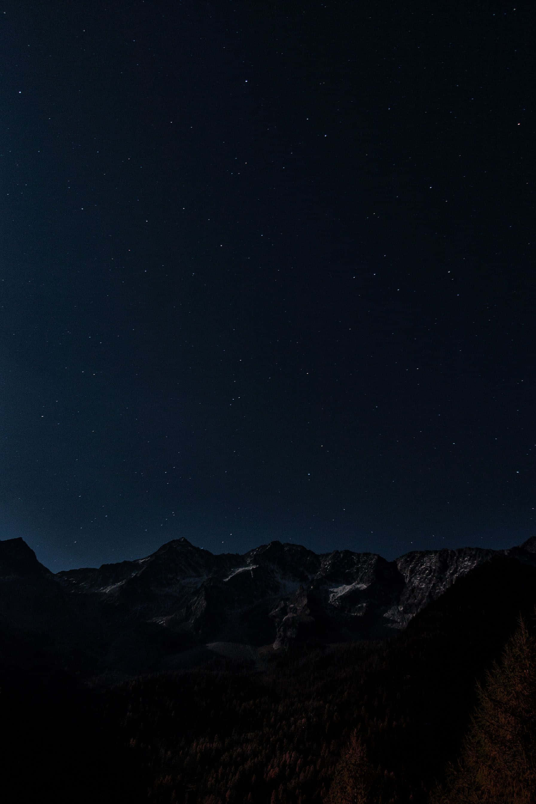 黑夜 桌布 壁紙 — 繁星點點的夜空拱形籠罩著黑暗的山脈，山頂覆蓋著白雪，下方是森林覆蓋的山坡。