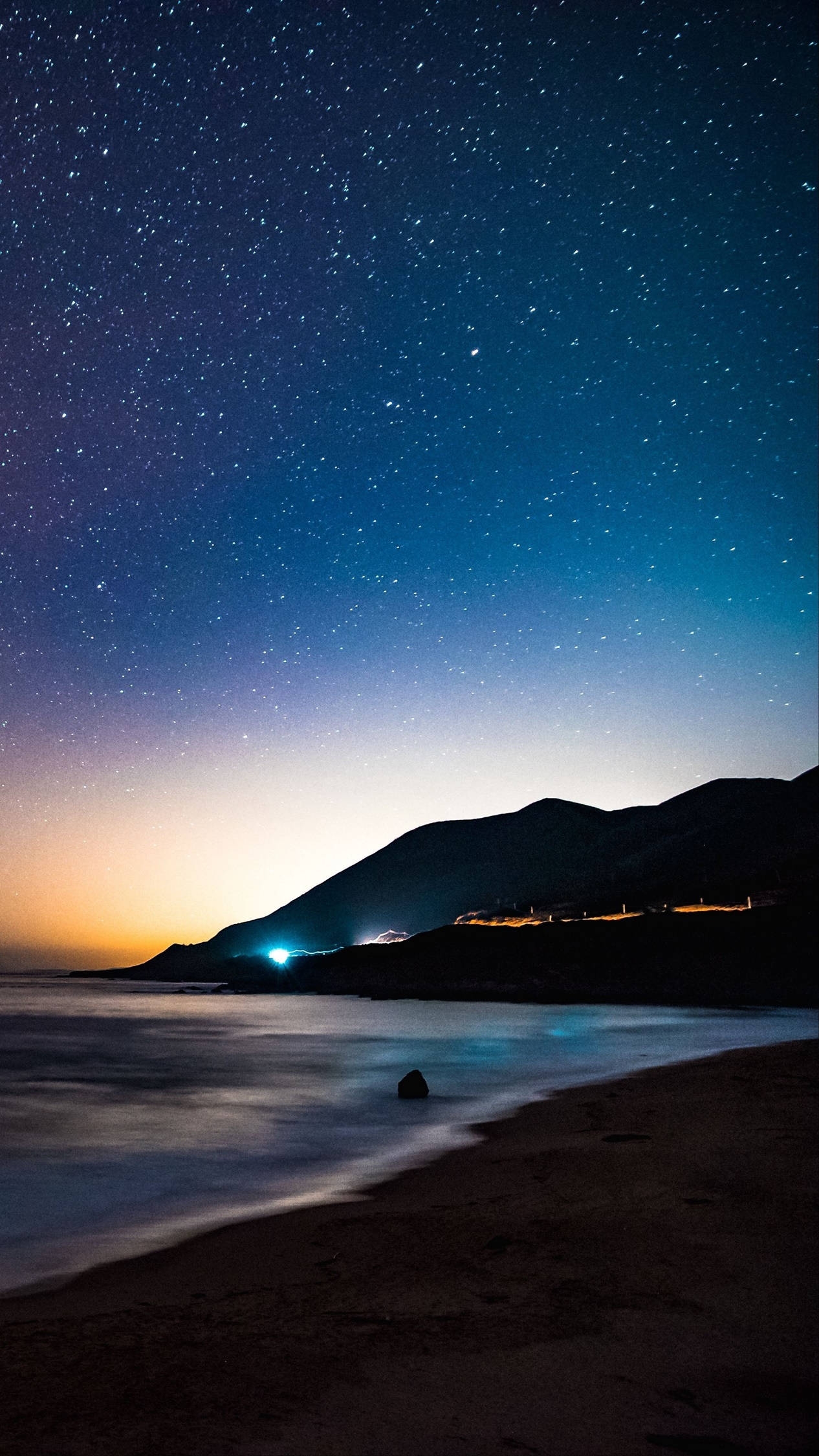 繁星點點的夜晚，海灘上有山丘和沿岸的遠方燈光。 — 4K Android 壁紙