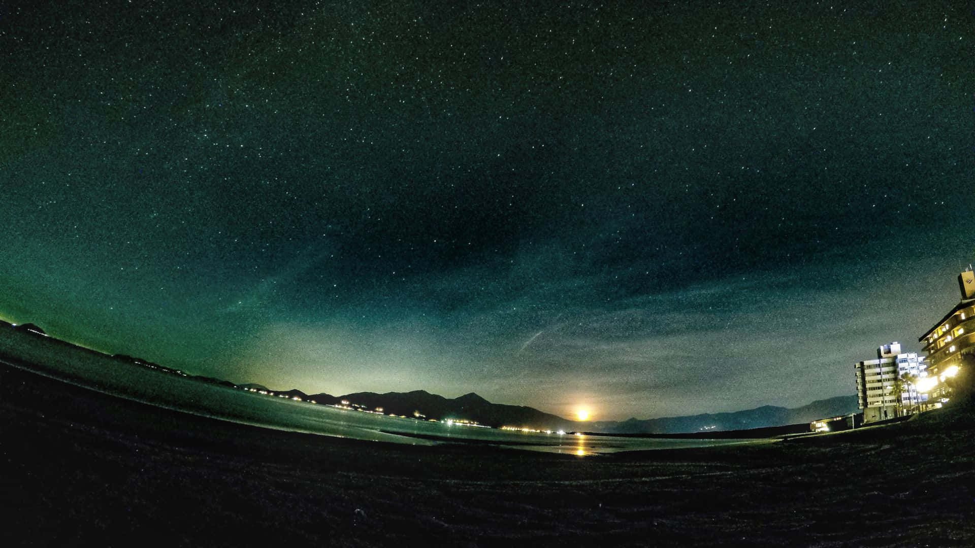 星空動畫 桌布 壁紙 — 繁星點點的夜空拱起在黑暗的海灘上，遠處有城市燈光和山脈，月亮閃耀。