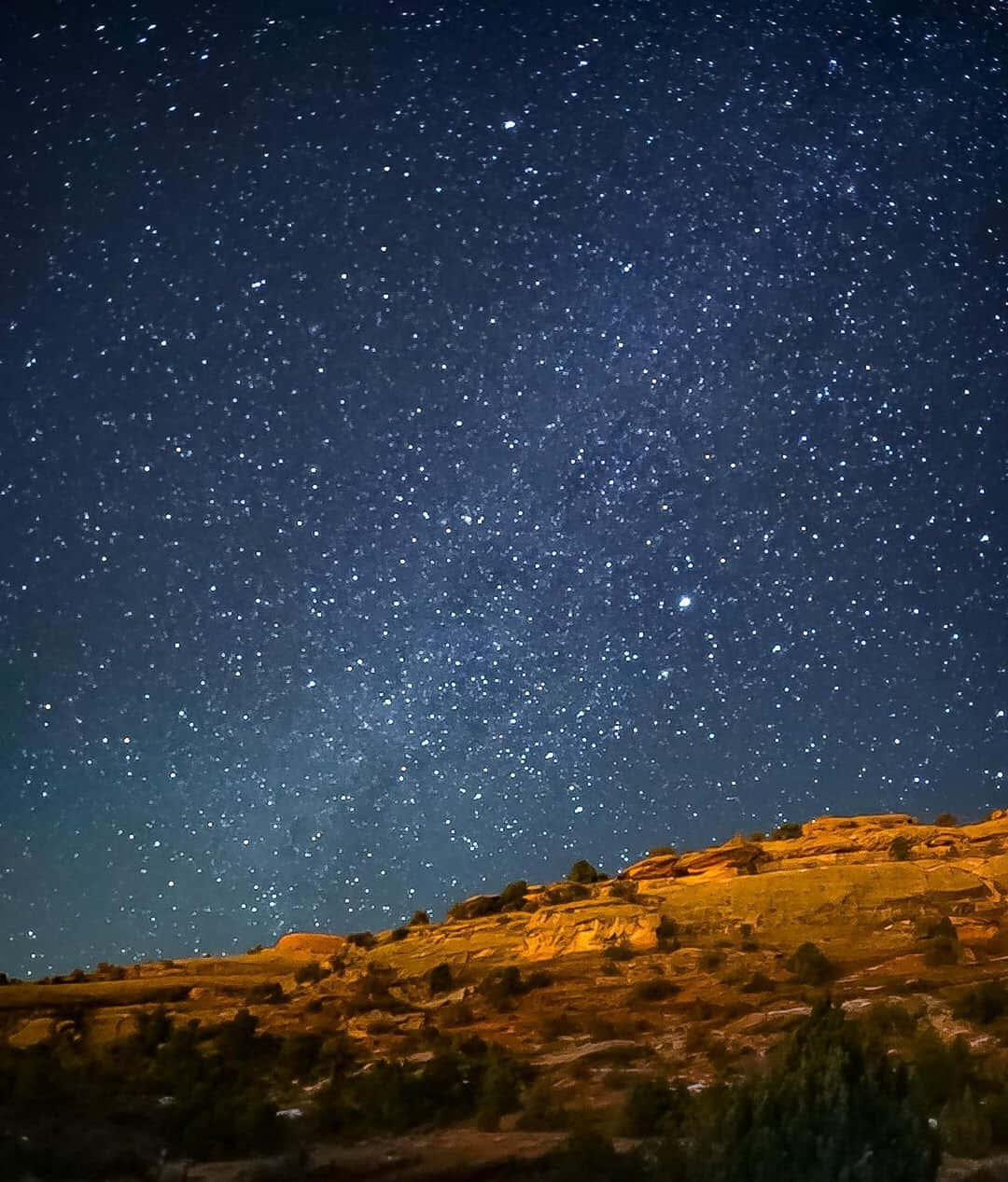 Starry Sky 桌布 壁紙 — 繁星點點的夜空覆蓋著崎嶇的沙漠山坡，散發著溫暖的人造光芒。