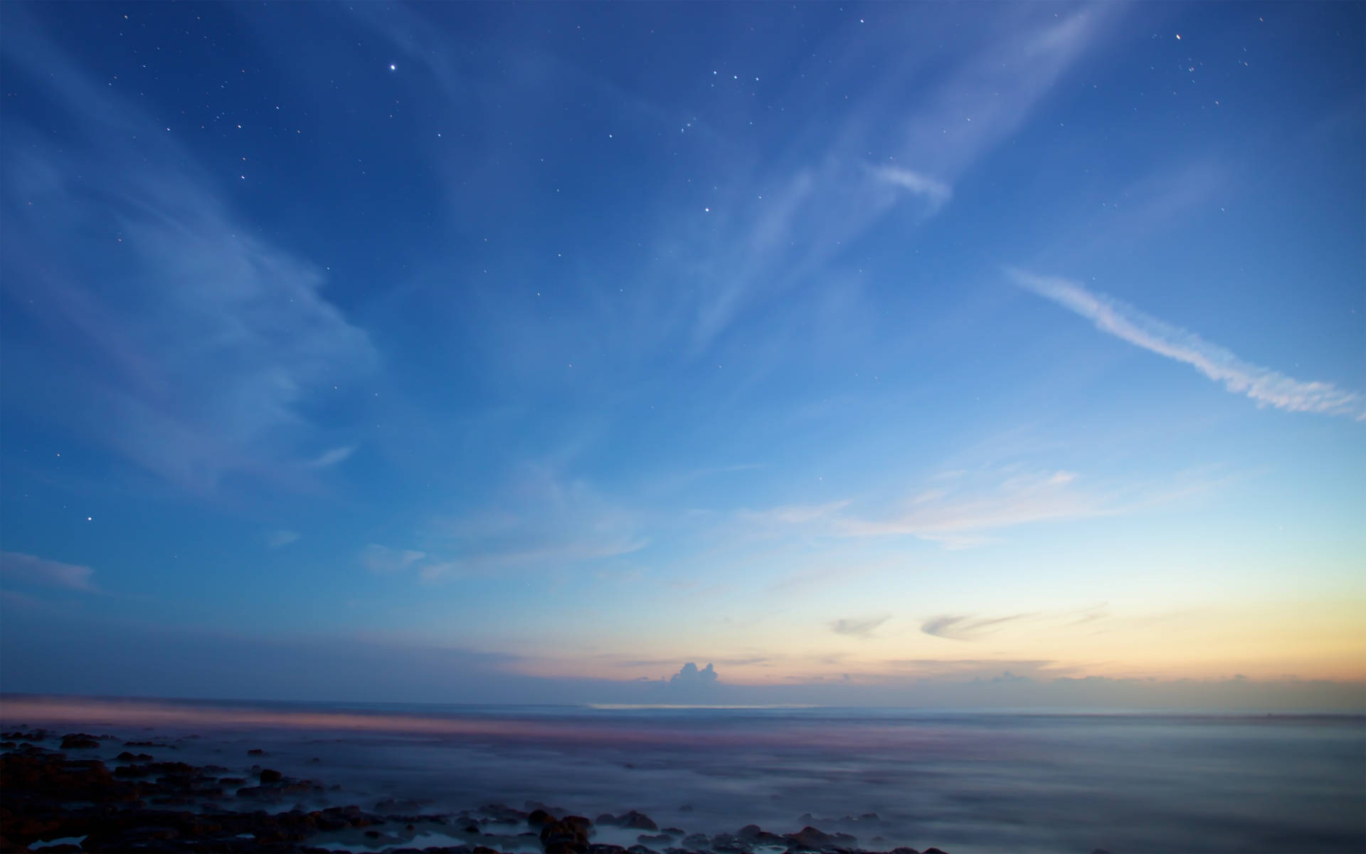 海洋 桌布 壁紙 — 繁星點點的天空中飄著縷縷薄雲，籠罩著暮色中平靜的海洋，前景是岩石海岸。