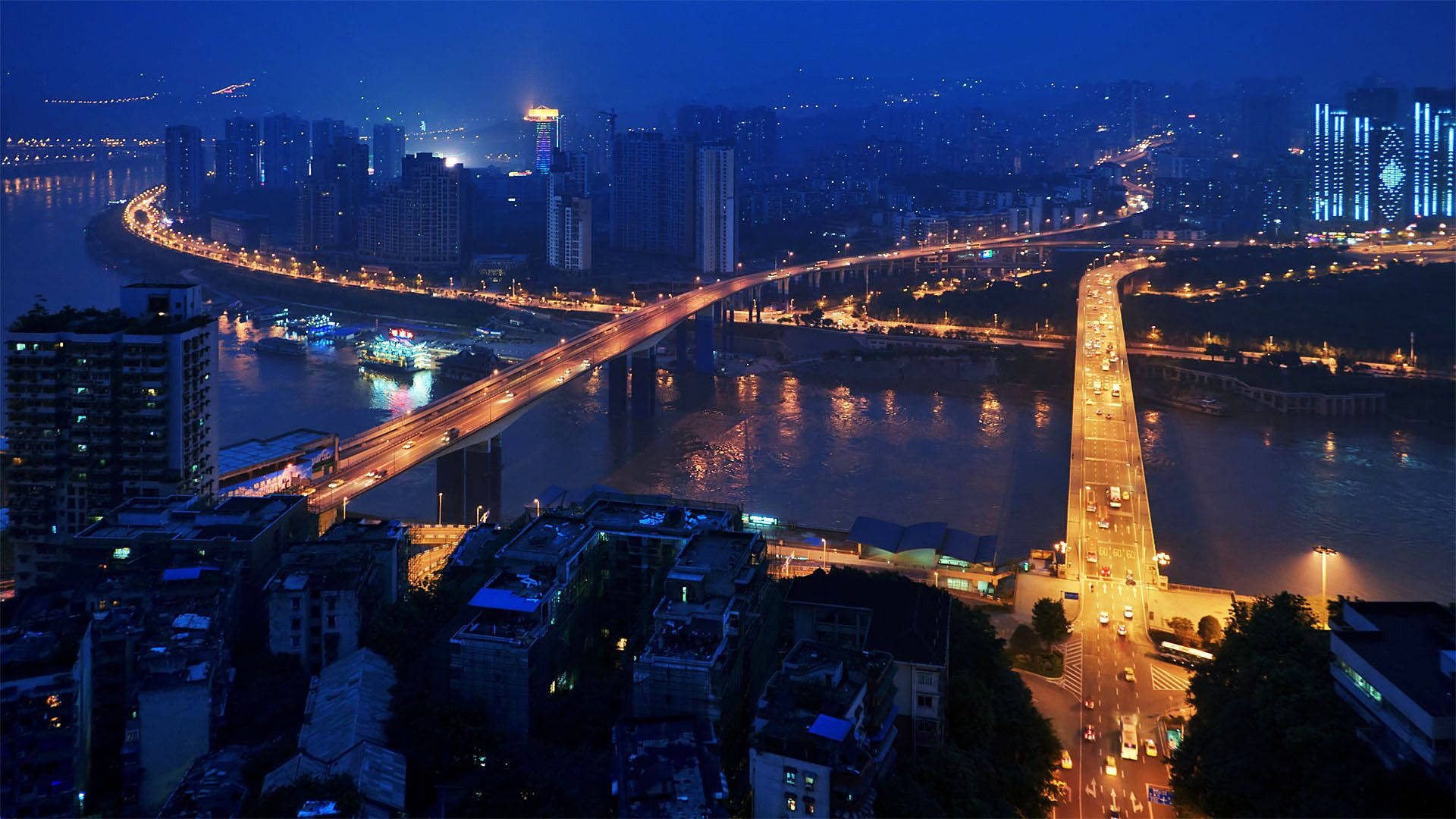 Chongqing 桌布 壁紙 — 繁華城市景觀的夜間鳥瞰圖，發光的橋樑和河流反射著充滿活力的街燈和摩天大樓。