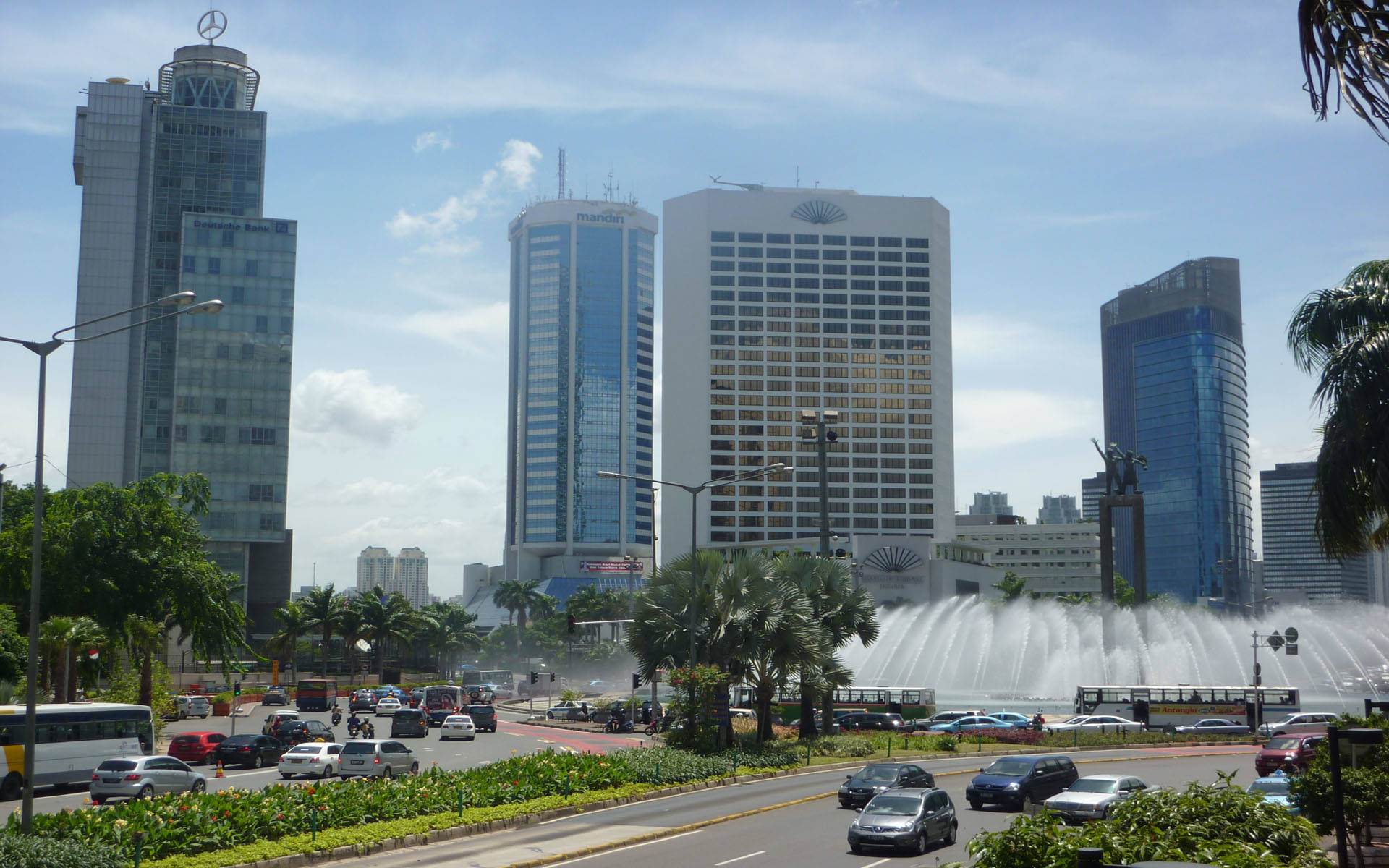 Jakarta 桌布 壁紙 — 繁華的城市景觀，擁有現代摩天大樓、棕櫚樹和一個在明亮藍天下噴水的大型噴泉。