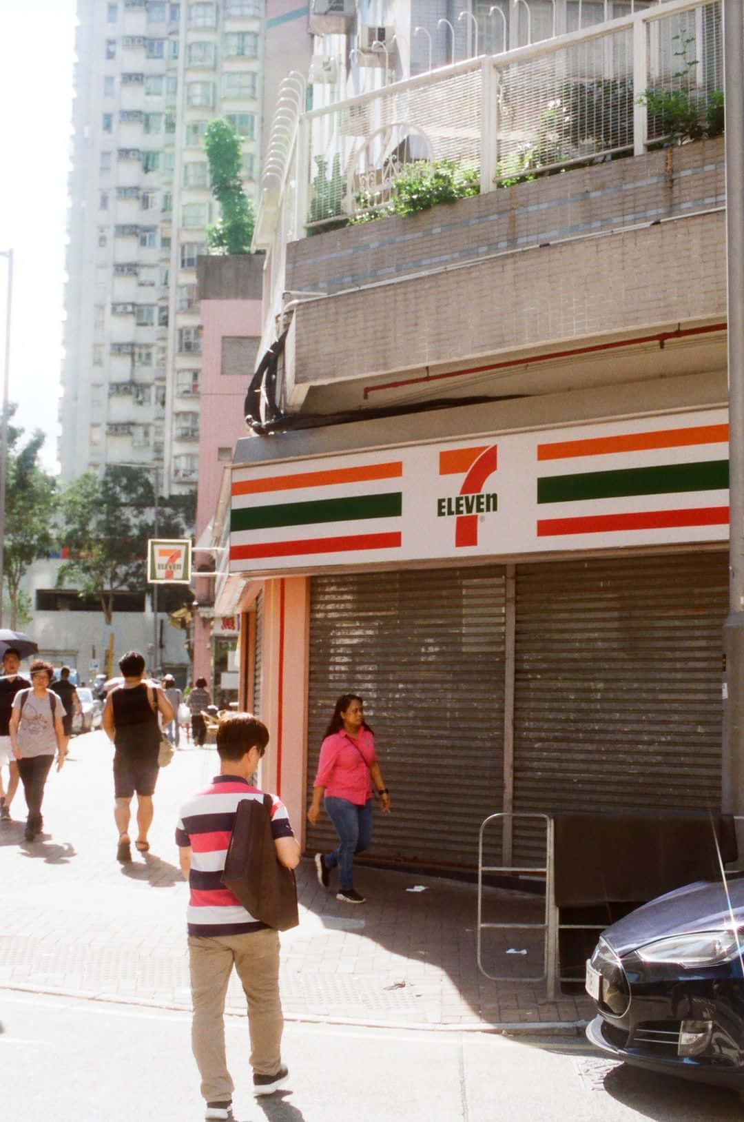 7-ELEVEN 桌布 壁紙 — 繁華的城市街景，行人經過一間關閉的7-Eleven商店，陽光普照。