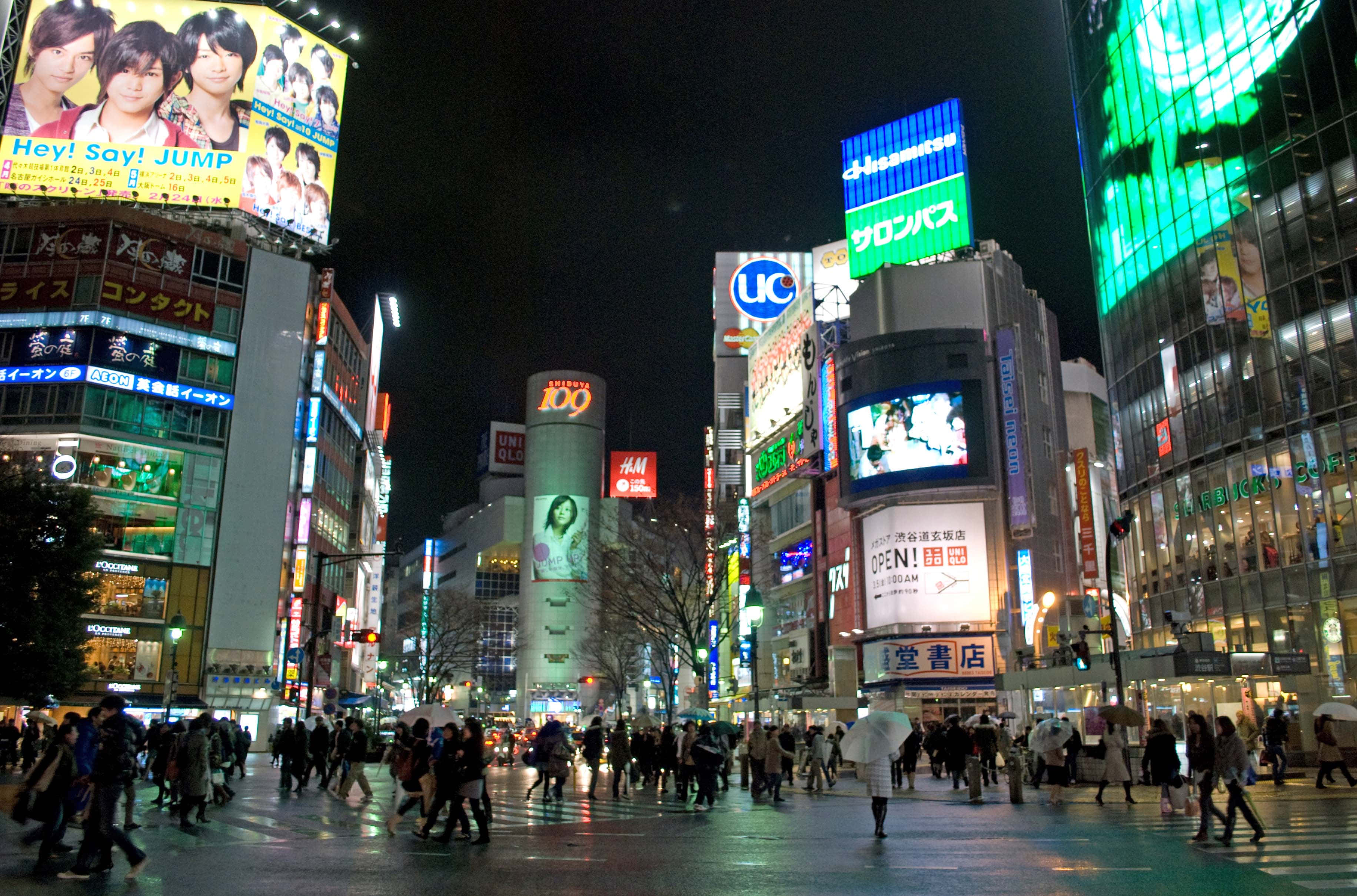 東京 桌布 壁紙 — 繁華的東京夜景，霓虹燈閃爍的摩天大樓、擁擠的街道，以及濕漉漉的人行道上反射的發光廣告牌。