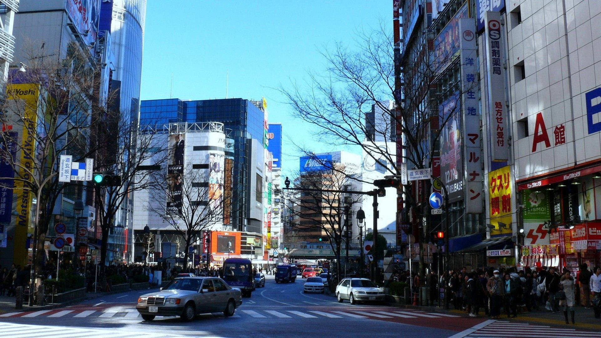 日本 桌布 壁紙 — 繁華的東京街道，高聳的建築物、霓虹燈招牌，行人在晴朗的藍天下過馬路。