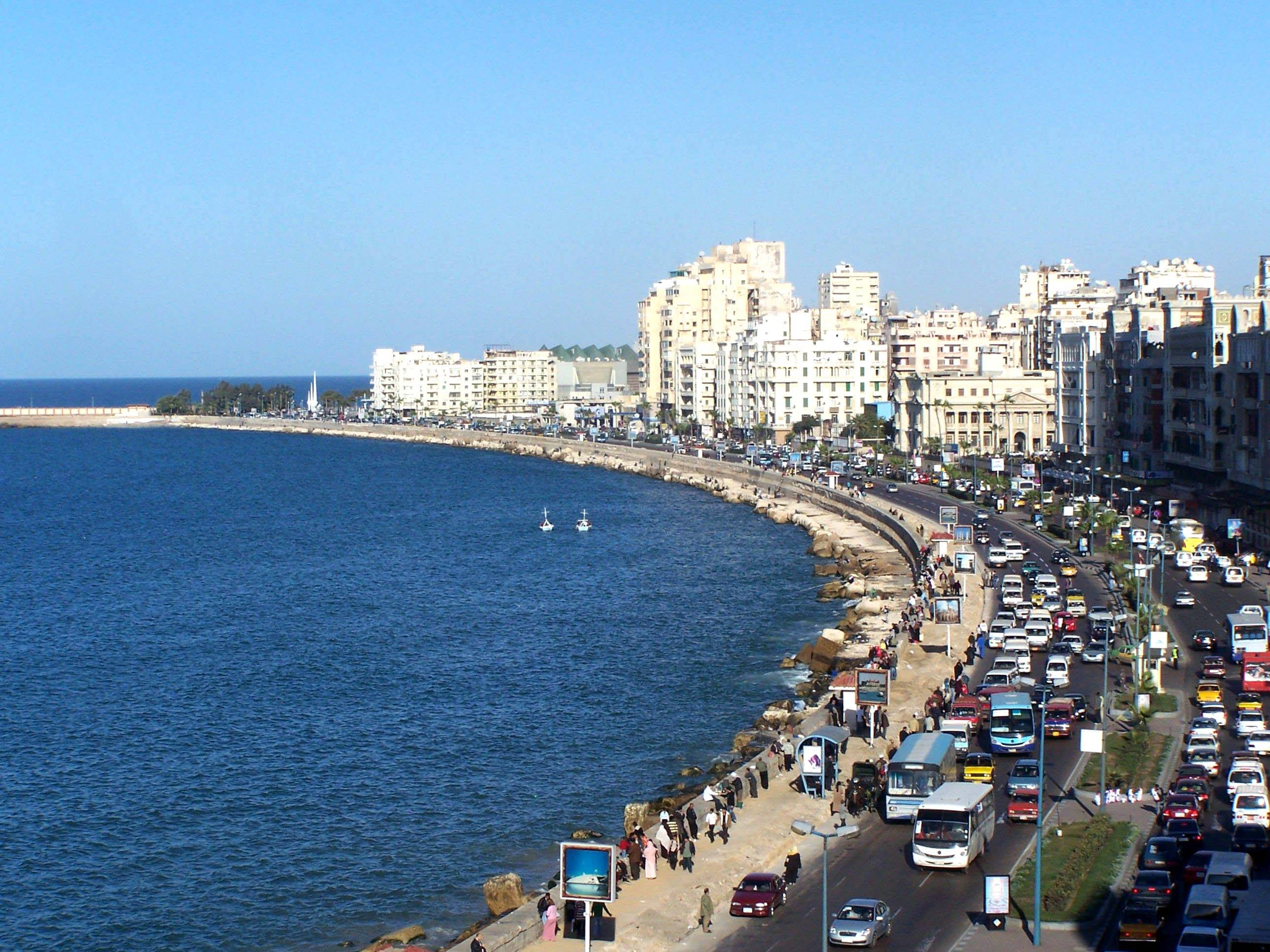 Alexandria 桌布 壁紙 — 繁華的海岸城市景觀，一條彎曲的道路車水馬龍，旁邊是深藍色的大海，天空晴朗。