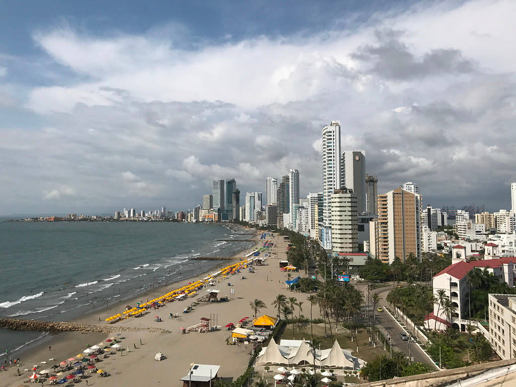 Colombia 桌布 壁紙 — 繁華的海濱城市景觀，高聳的摩天大樓沿著沙灘排列，沙灘上佈滿色彩繽紛的遮陽傘和棕櫚樹，天空多雲。