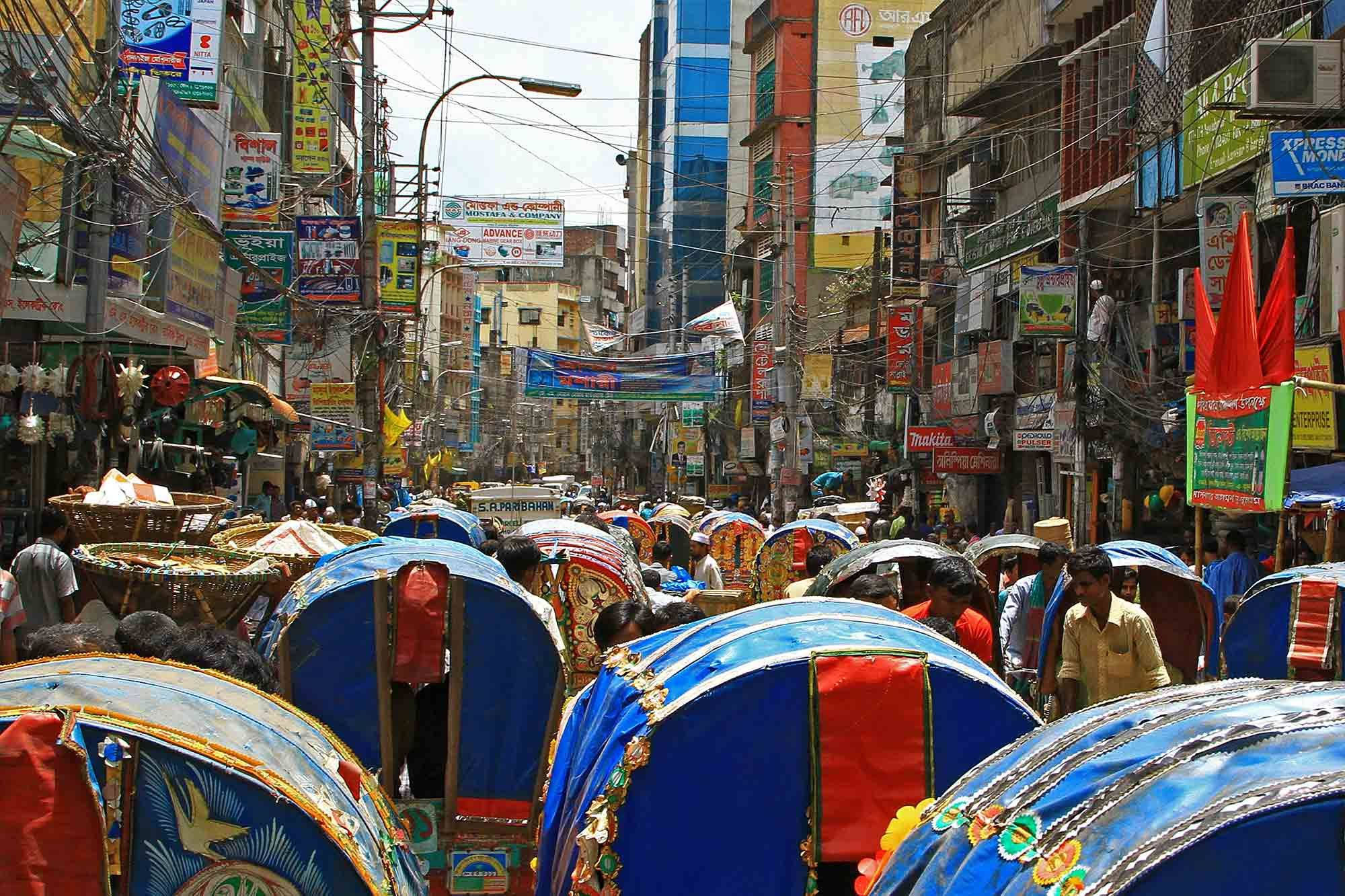 Dhaka 桌布 壁紙 — 繁華的都市街道，色彩繽紛的人力車和擁擠的商店，天空明亮。