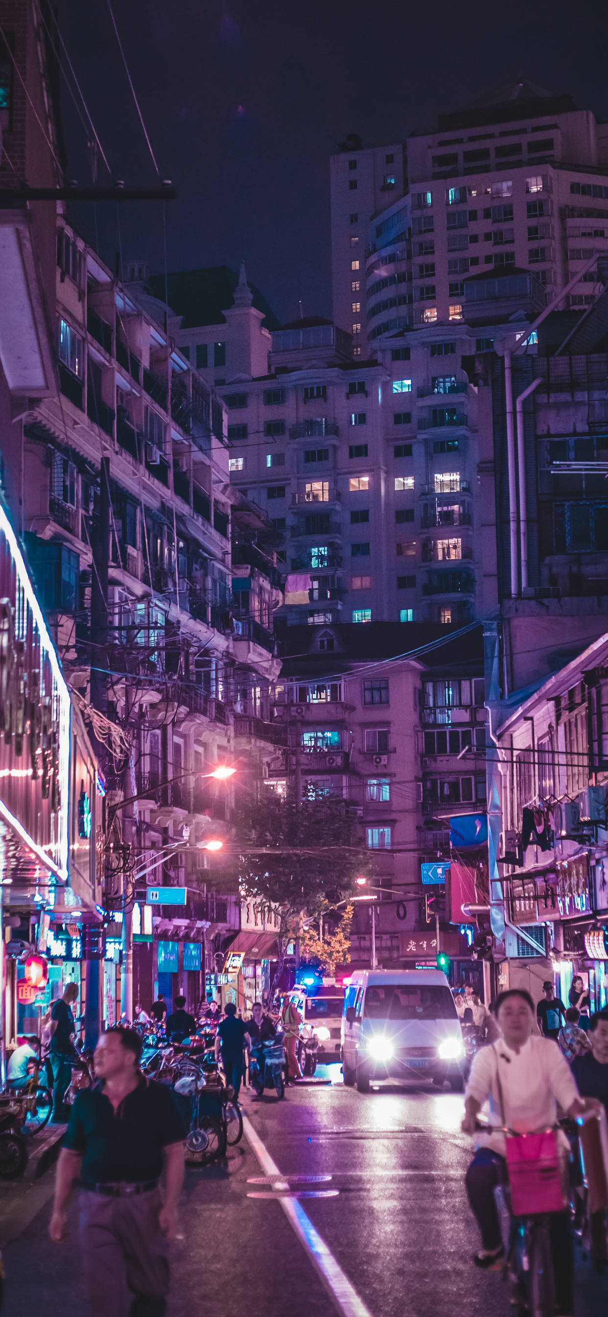 城市 桌布 壁紙 — 繁華的霓虹燈照亮城市街道夜晚，行人、自行車騎士和發光的商店櫥窗在高聳的公寓大樓之間。