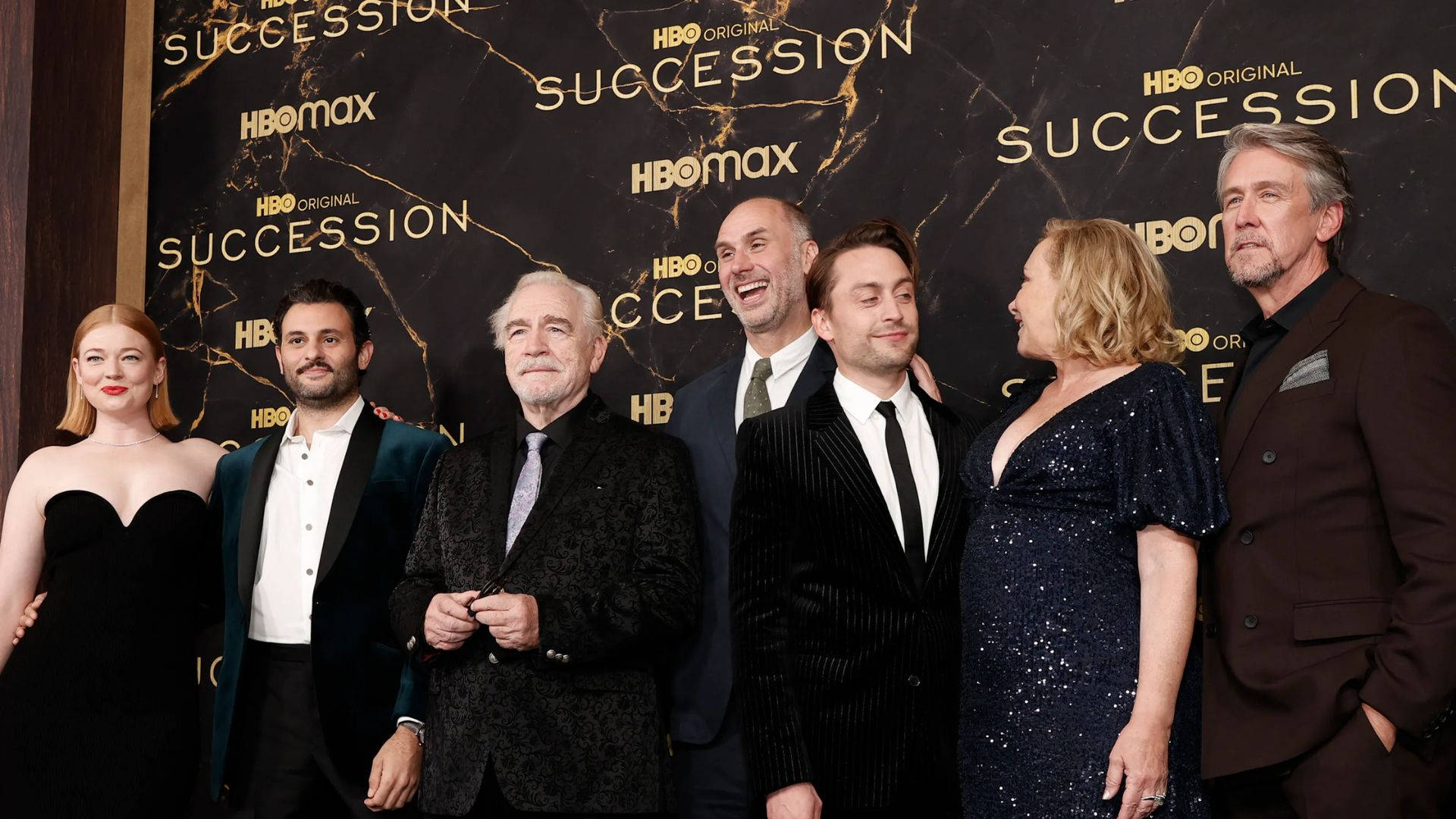 Succession 桌布 壁紙 — 《繼承之戰》演員在紅毯活動中正式擺姿勢，背景為黑色大理石配金色文字。