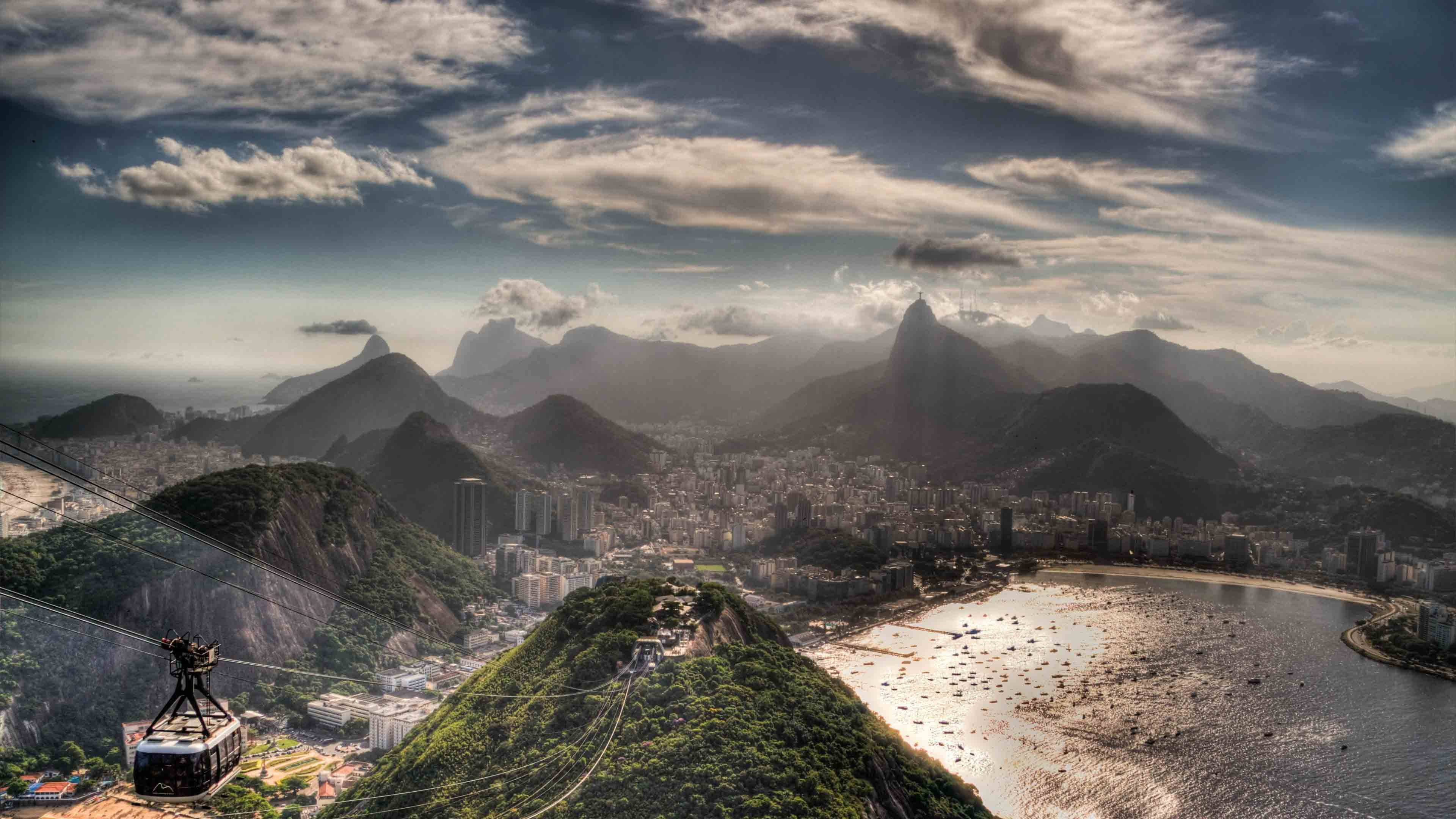 South America 桌布 壁紙 — 纜車在戲劇性的多雲天空下滑過鬱鬱蔥蔥的綠色山脈，俯瞰里約熱內盧廣闊的城市景觀和閃閃發光的海灣。