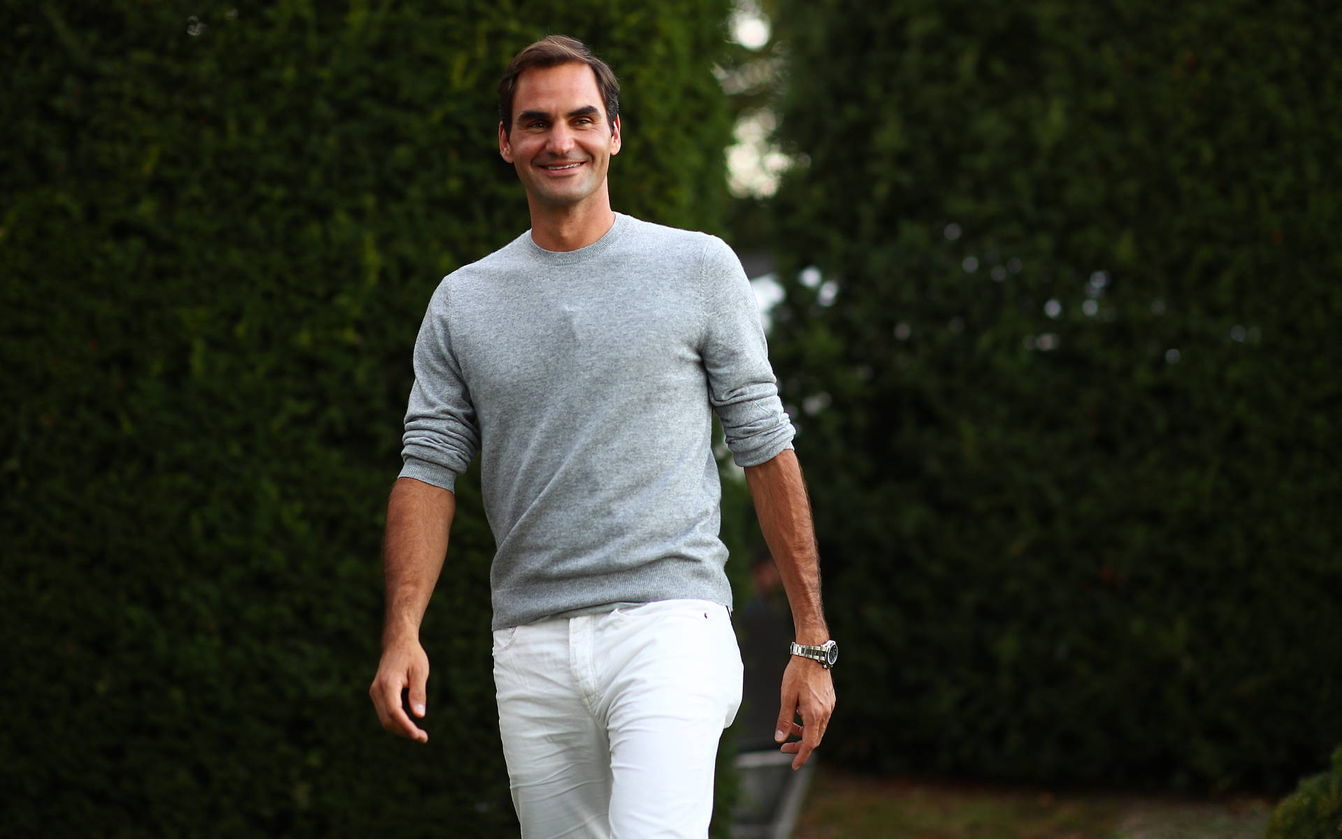 roger 桌布 壁紙 — 羅傑·費德勒穿著灰色毛衣和白色長褲，在茂密的綠色樹籬背景下自信地行走。