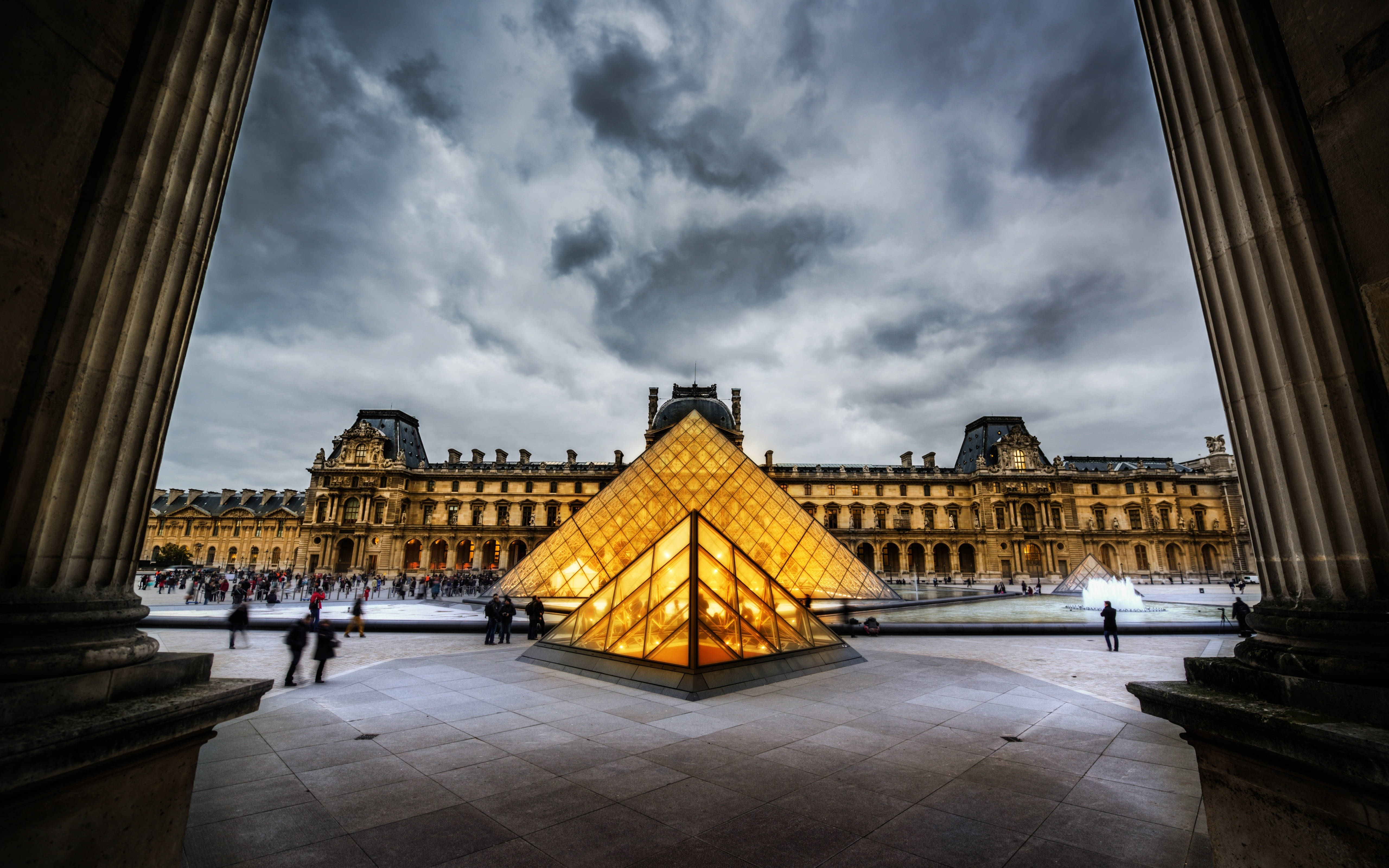 France 桌布 壁紙 — 羅浮宮博物館中發光的玻璃金字塔，在戲劇性的多雲天空下被宏偉的柱子環繞。